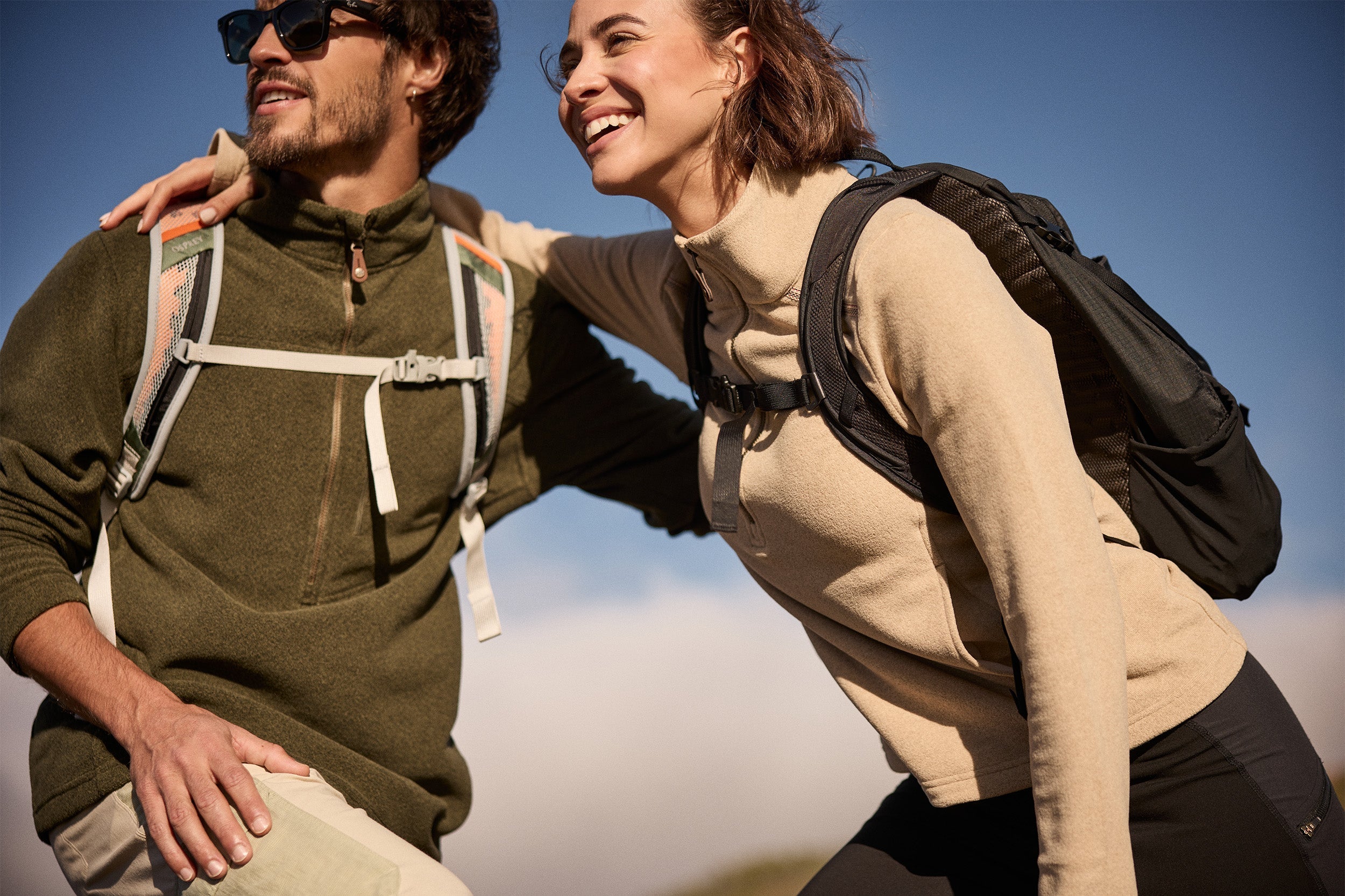 A man and woman standing at the top of a hill. The man is wearing a green fleece and black sunglasses. The women is wearing a beige fleece and black leggings.