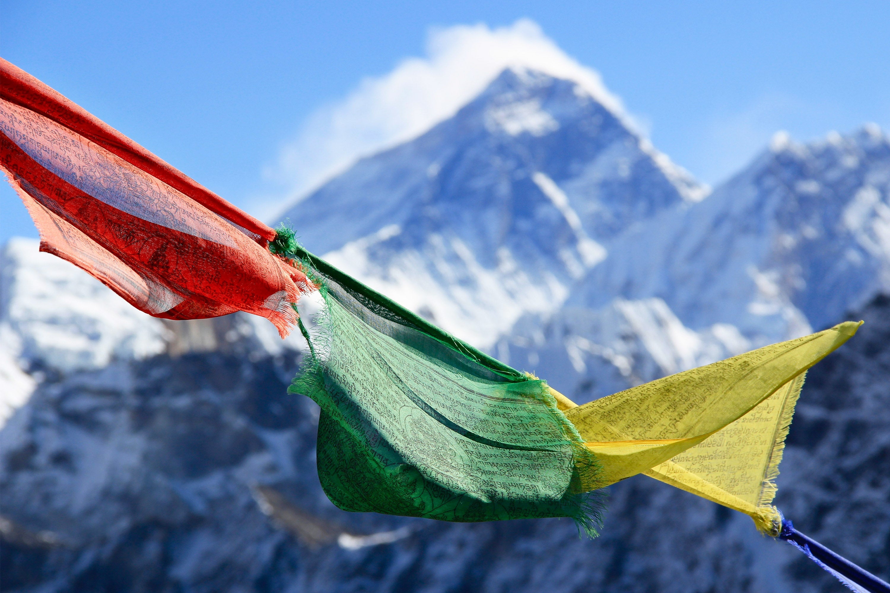 A row of red, green and yellow prayer flags sway in the breeze in front of Mount Everest in Nepal. The mountain is covered in snow with a blue sky behind.