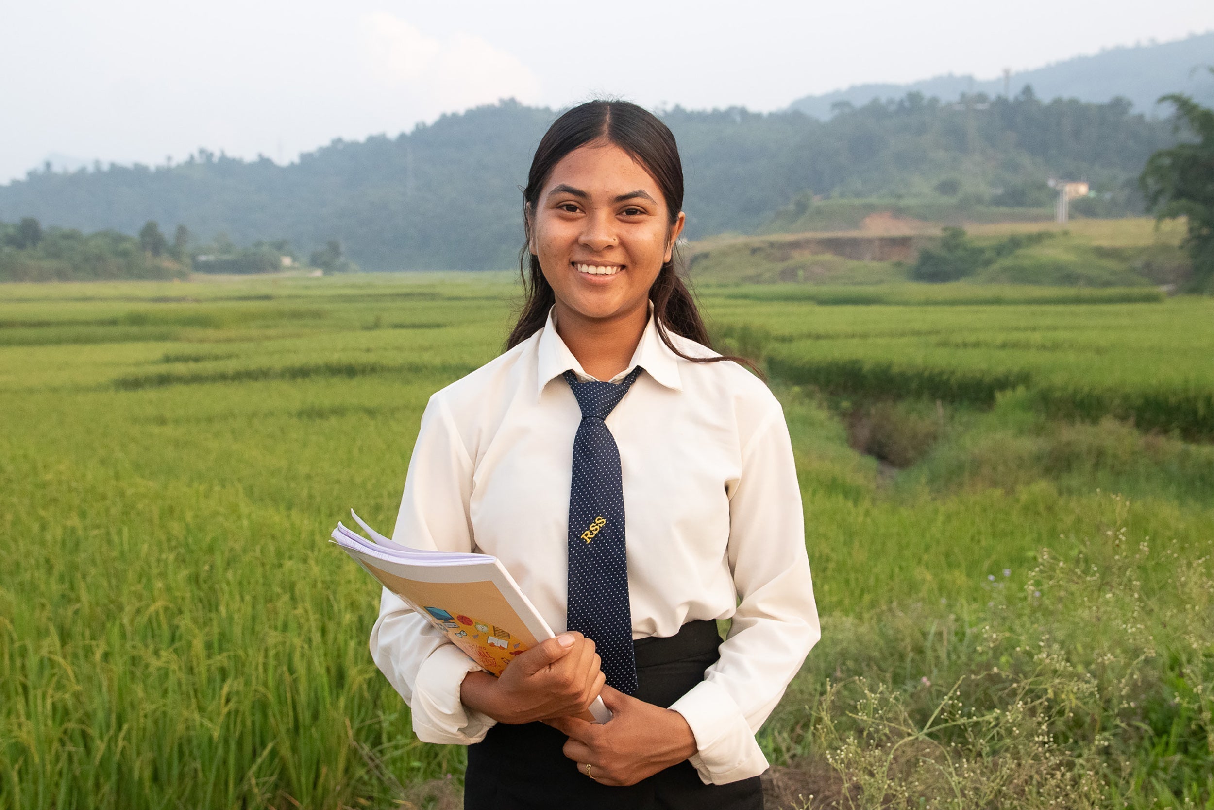 Sulochana smiling in white shirt, blue tie, holding open colorful notebook