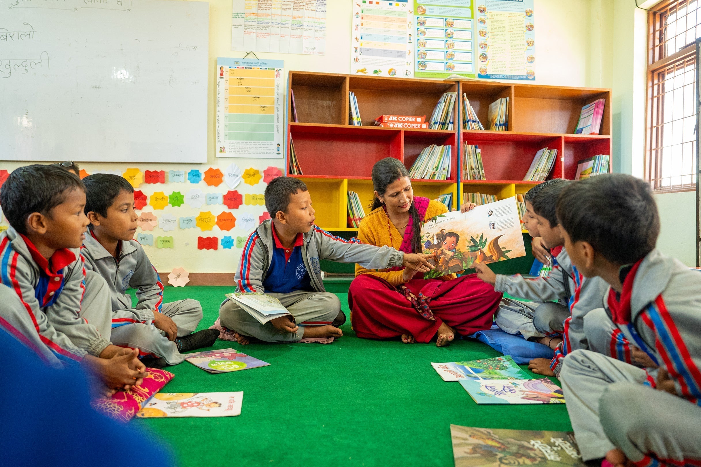 Woman in orange top and red pants reads kids book on green floor for Give Back Tuesday