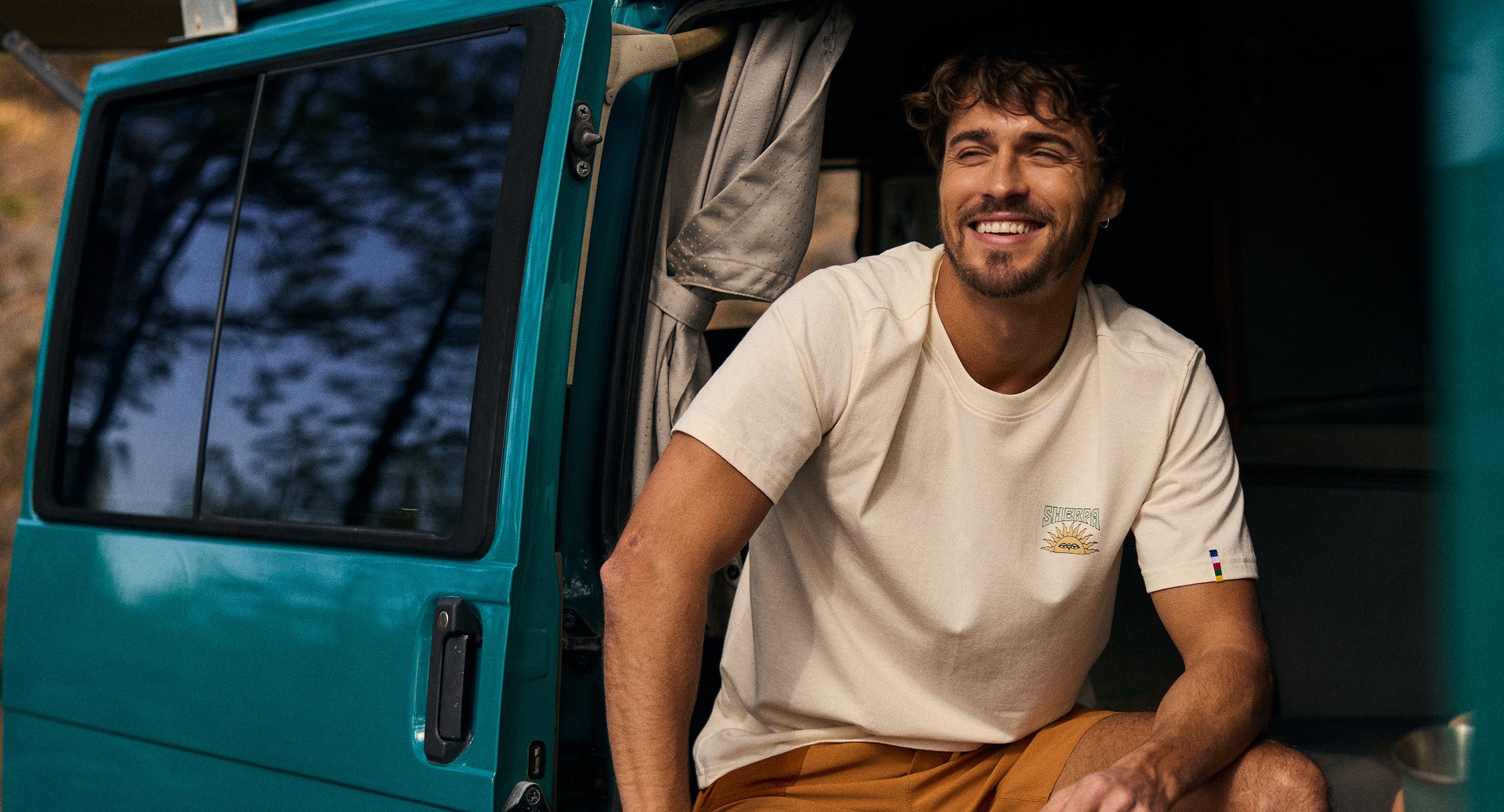 Man sitting inside a teal van holding a mug, wearing a beige t-shirt and orange shorts.