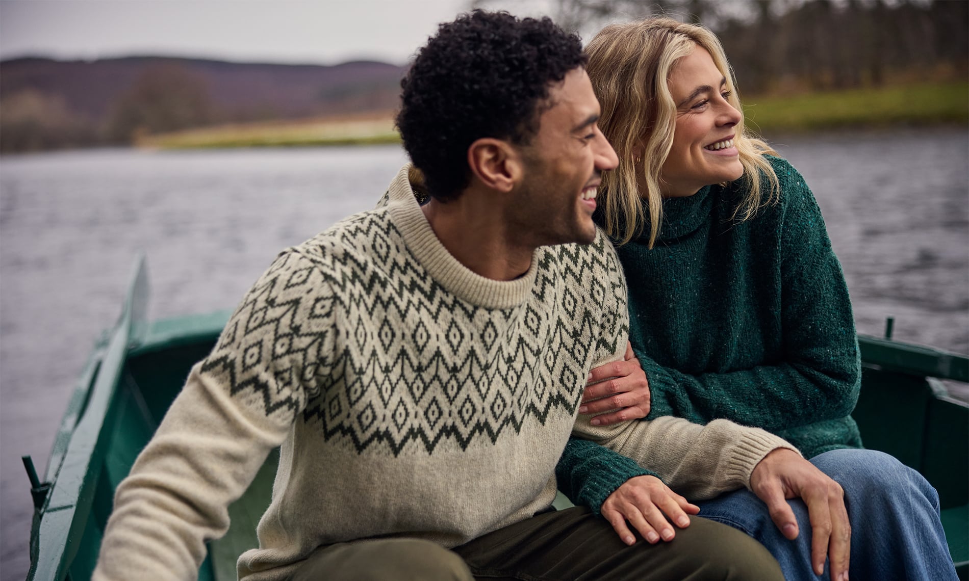 A man and a woman sitting together in a green boat on a lake, wearing cozy knit sweaters. The man wears a cream sweater with a dark geometric pattern, and the woman wears a dark green sweater.