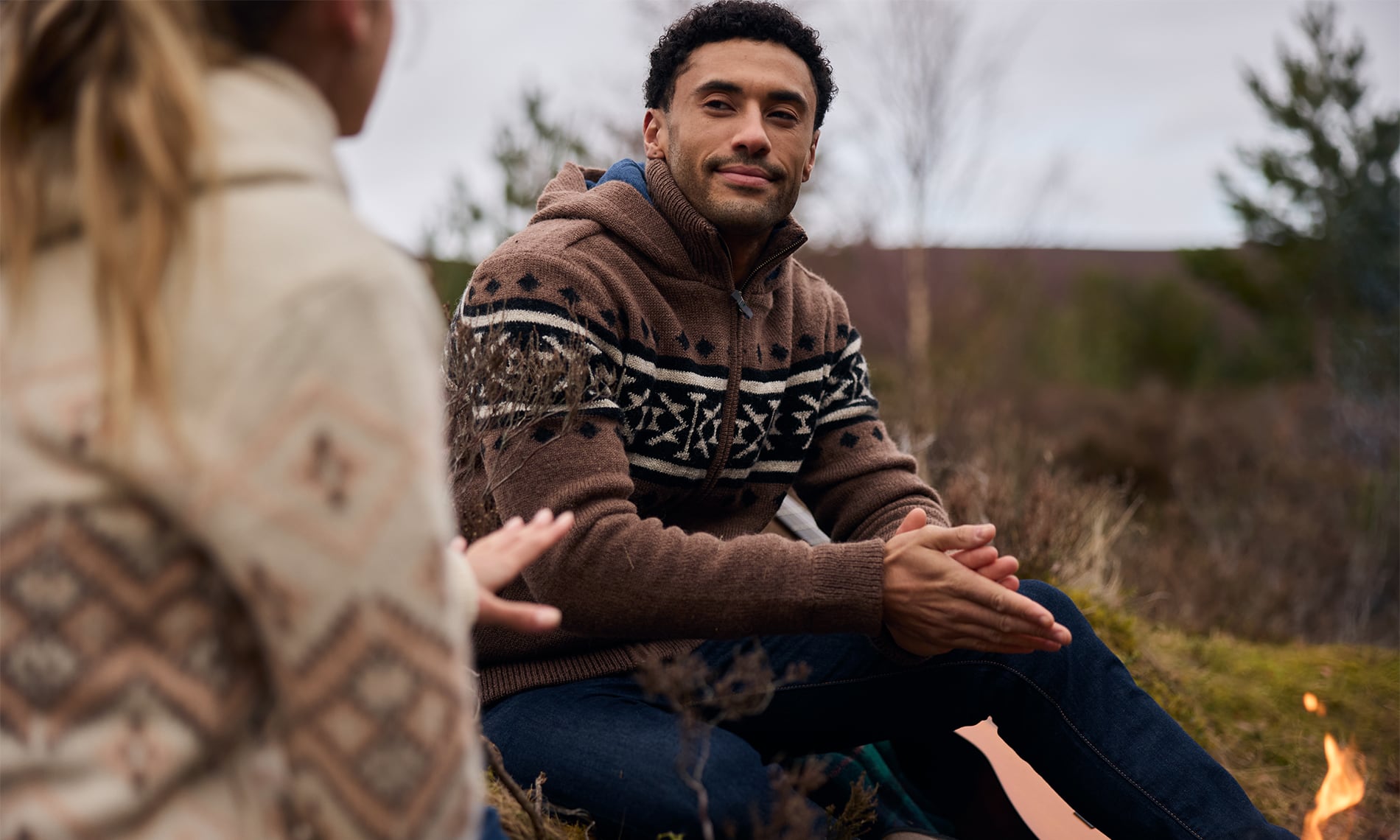A man wearing a brown knit zip-up hoodie with a white and brown Nepal inspired pattern sits outdoors near a campfire, smiling at a woman in a matching patterned sweater.