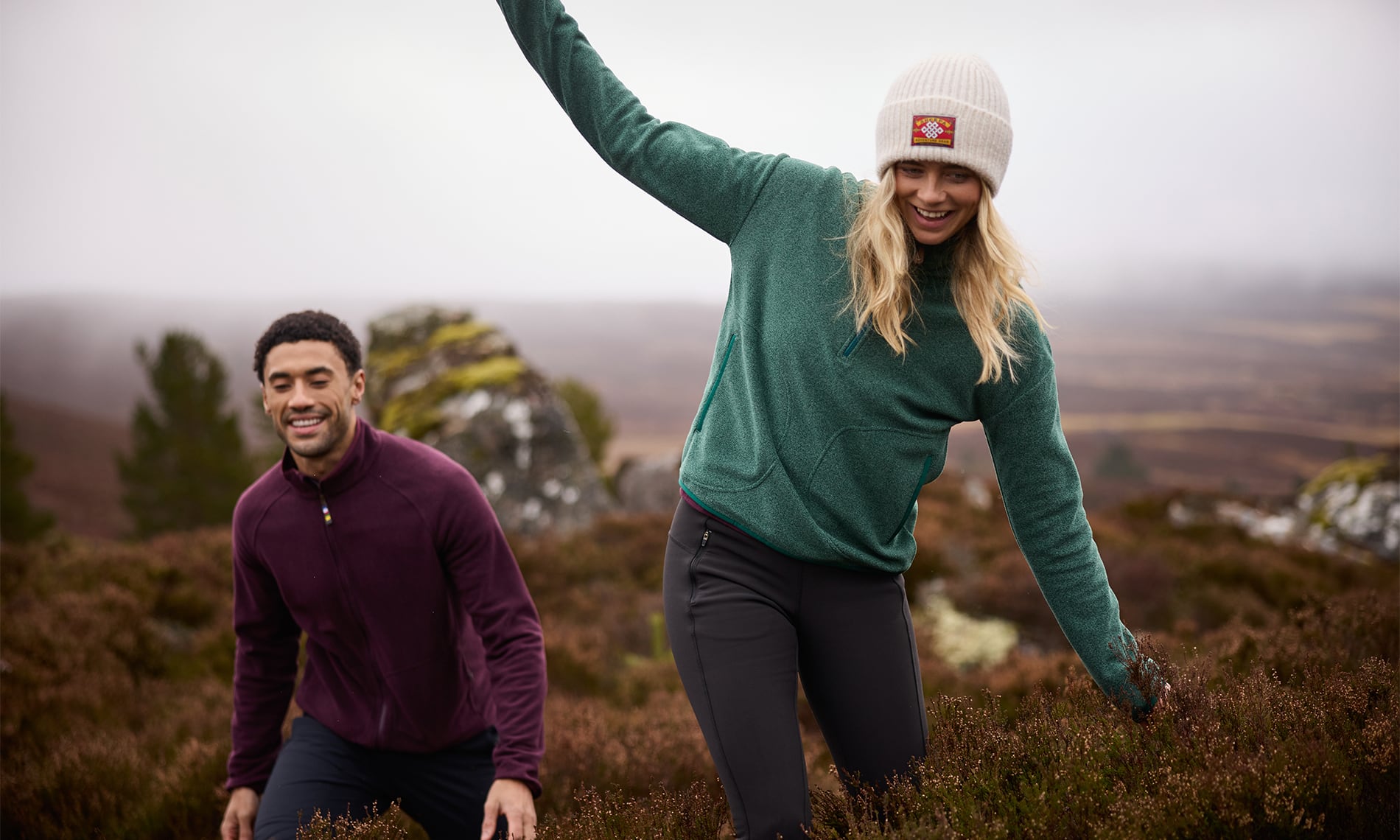 A man and a woman outdoors in a misty, hilly landscape wearing matching knit quarter-zip fleece pullovers. The woman wears a teal-green pullover and a white beanie with a small red square logo patch, while the man wears a dark plum-purple fleece jacket.