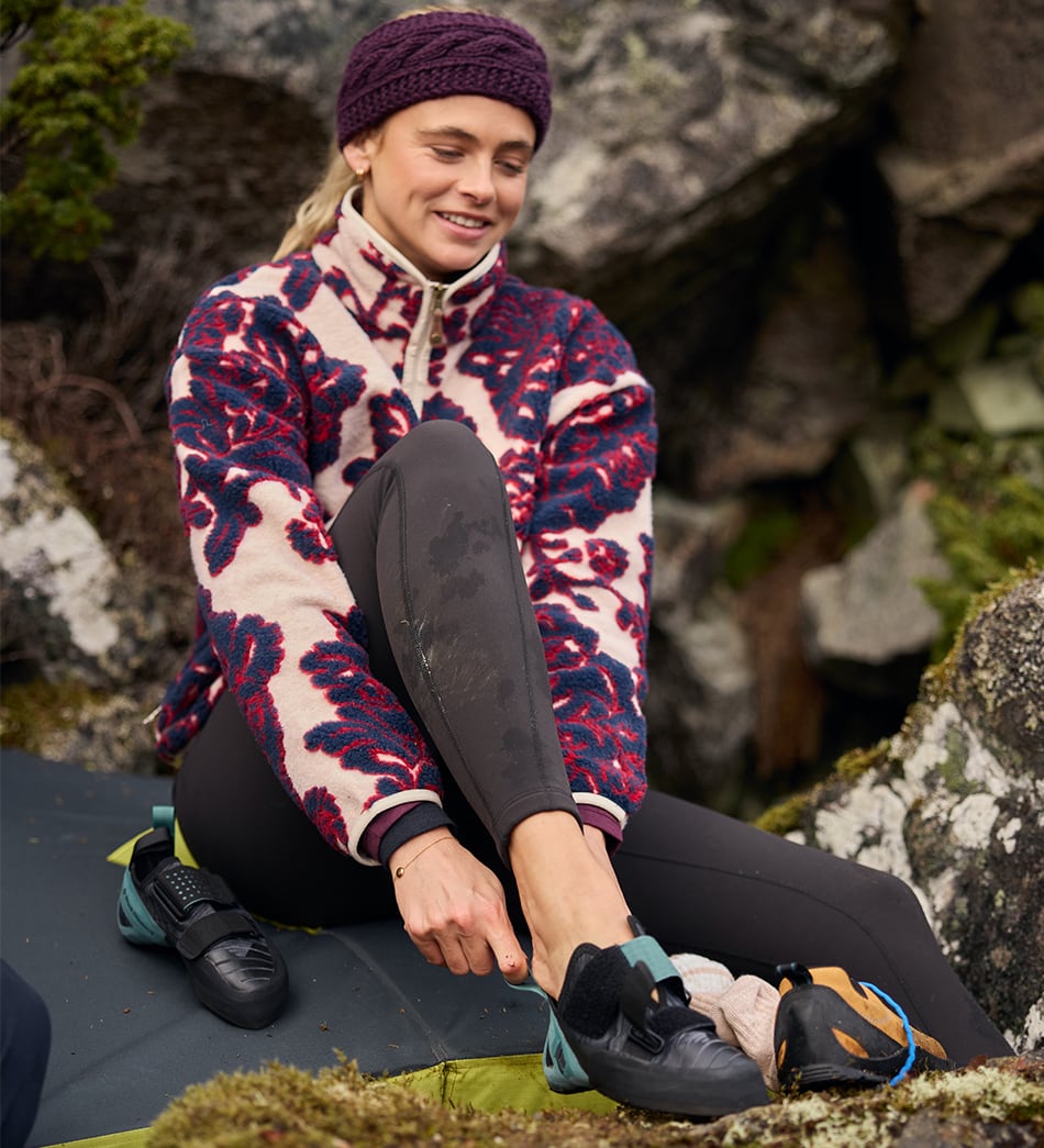 A woman sitting outdoors on a rock among mossy stones, wearing a cream fleece jacket with a red and navy floral pattern, black climbing pants, pulling on a pair of climbing shoes.