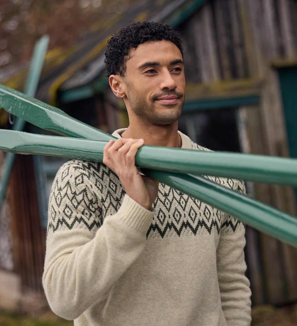 A man wearing a cream-colored knit sweater with a black geometric pattern across the chest, holding a green wooden rowing oars outdoors in front of a rustic wooden building.