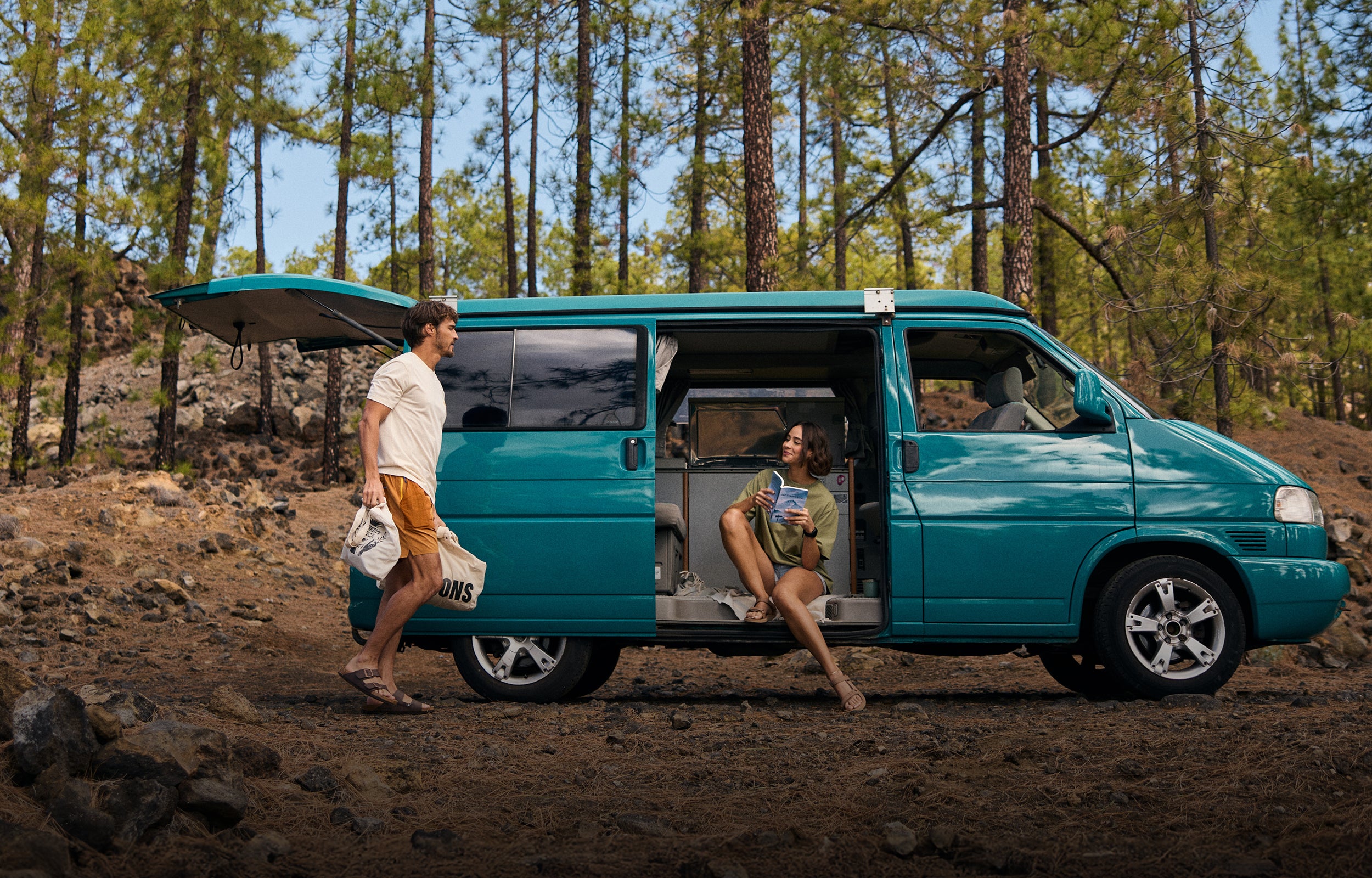 A man and woman by a teal camper van in a forest setting. The man walking by the side of the van wearing a white T-Shirt and a pair of yellow shorts. The woman is sat in the van wearing a green T-Shirt and pale blue pair of shorts.