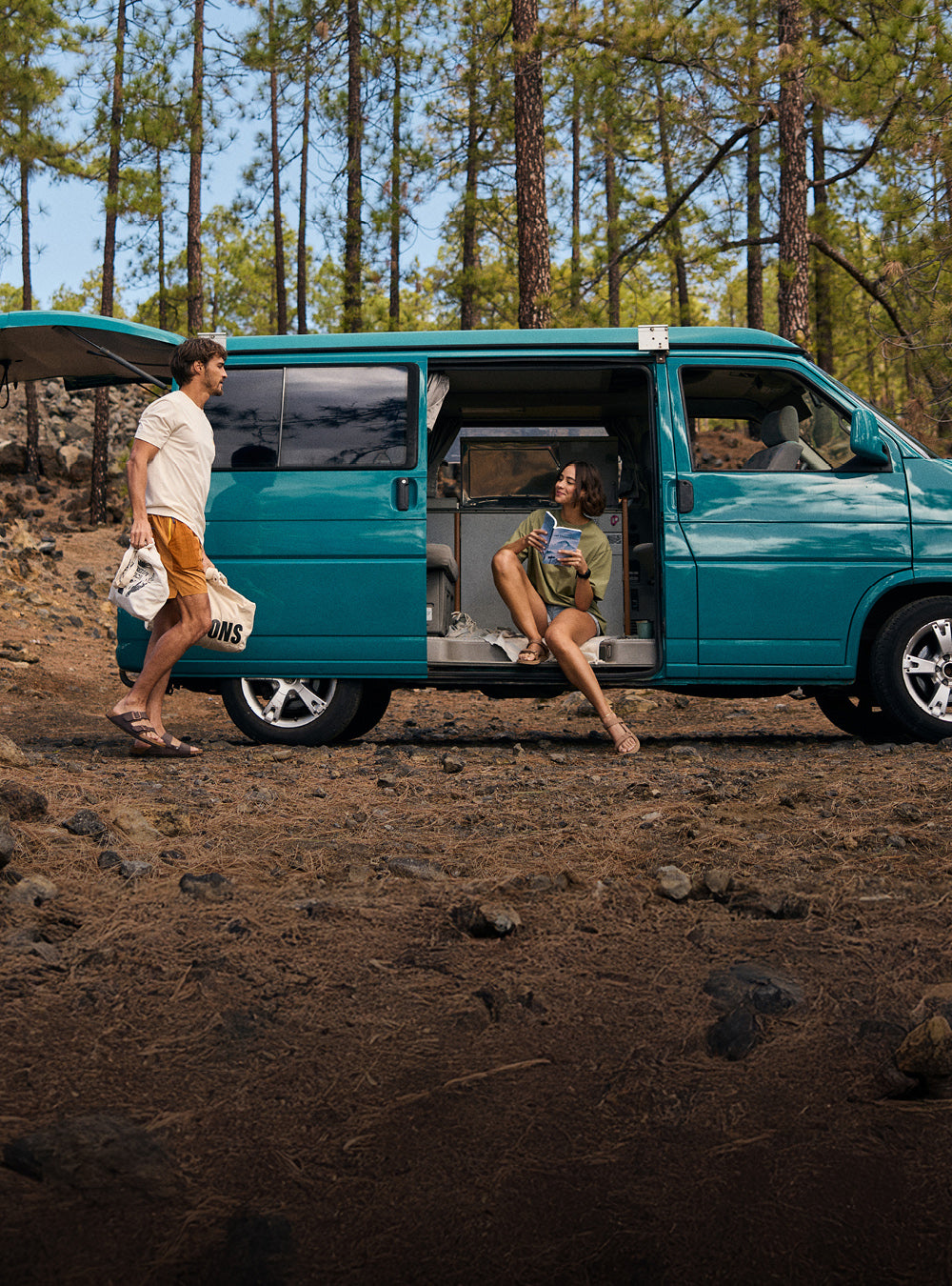 A man and woman by a teal camper van in a forest setting. The man walking by the side of the van wearing a white T-Shirt and a pair of yellow shorts. The woman is sat in the van wearing a green T-Shirt and pale blue pair of shorts.