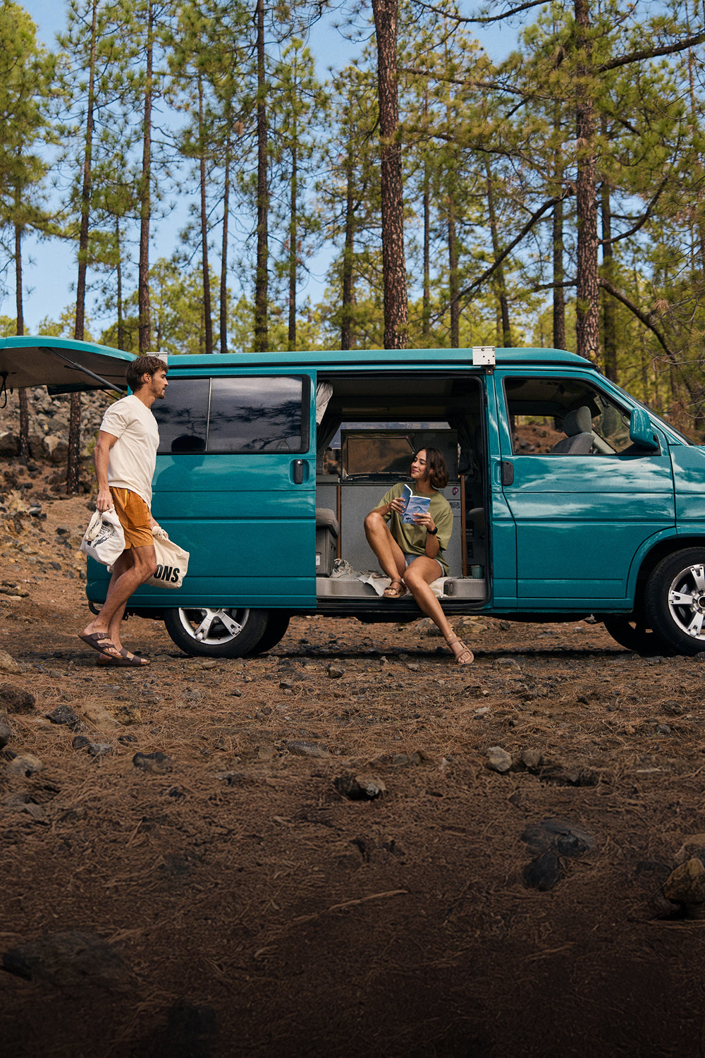 A man and woman by a teal camper van in a forest setting. The man walking by the side of the van wearing a white T-Shirt and a pair of yellow shorts. The woman is sat in the van wearing a green T-Shirt and pale blue pair of shorts.