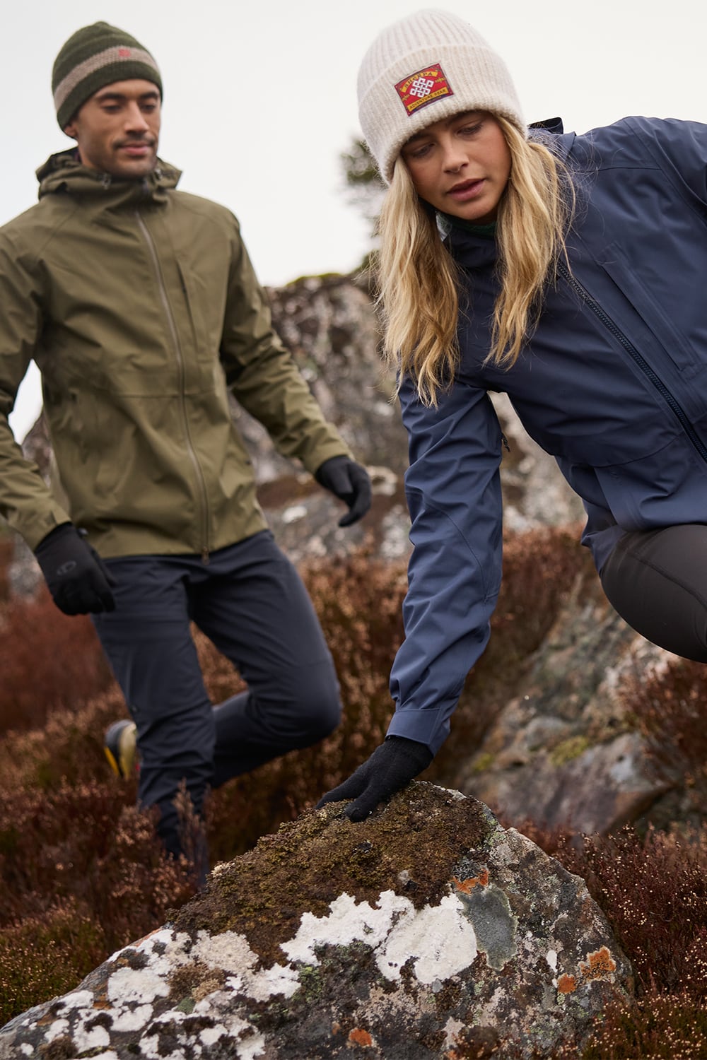 Two outdoor adventurers hiking on a rocky, mossy mountain path. The woman in the foreground wears a navy blue jacket, a white knit beanie with a red logo, black gloves, and black leggings. The man in the background wear a green waterproof Sherpa Adventure Gear jacket with a matching green beanie.