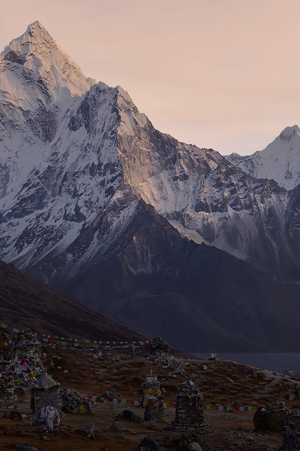 A wide view of snow-covered Himalayan mountains at sunrise, with the sun illuminating the peaks. Darker, rocker mountains sit in the foreground with colourful prayer flags hanging on the lower hills.