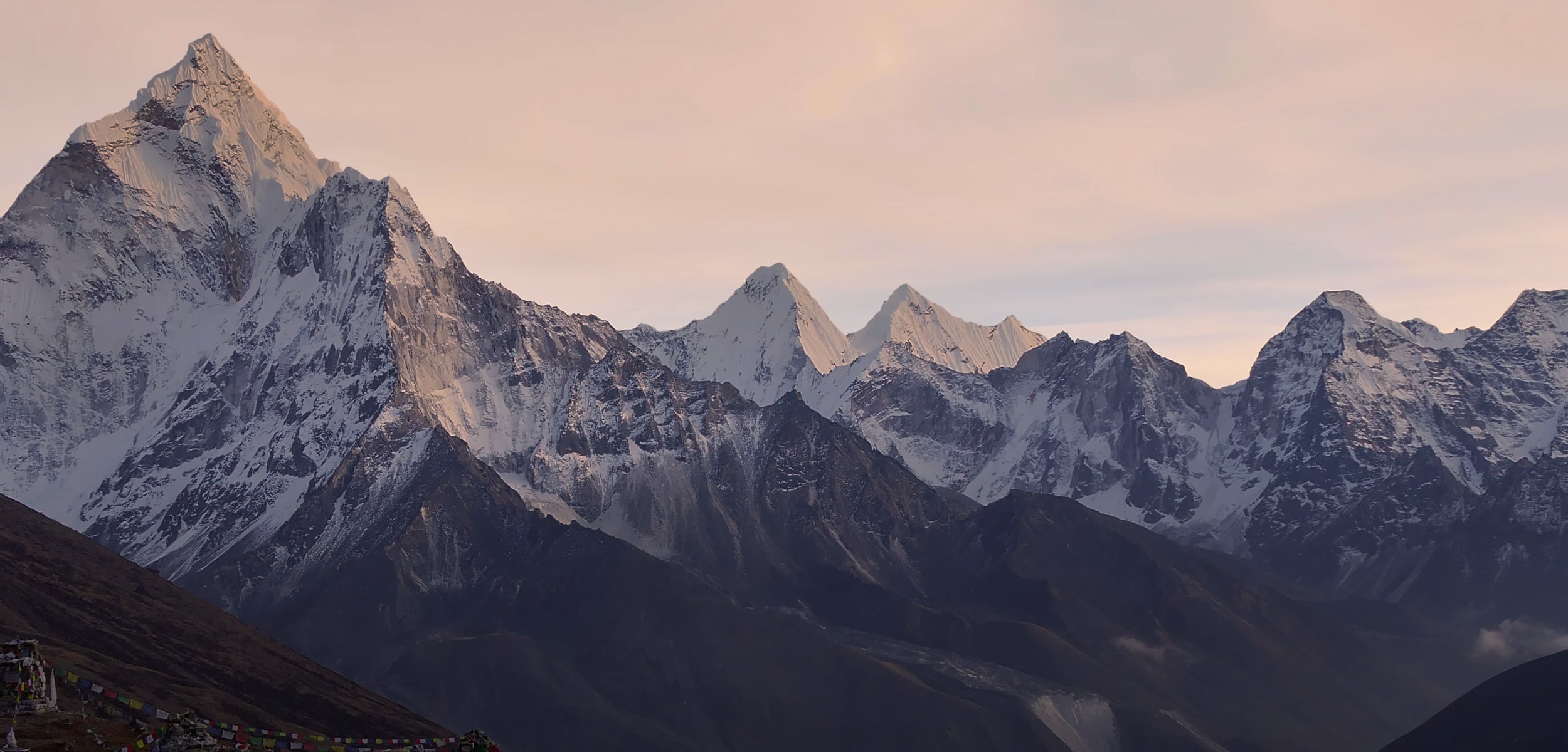A wide view of snow-covered Himalayan mountains at sunrise, with the sun illuminating the peaks. Darker, rocker mountains sit in the foreground with colourful prayer flags hanging on the lower hills.