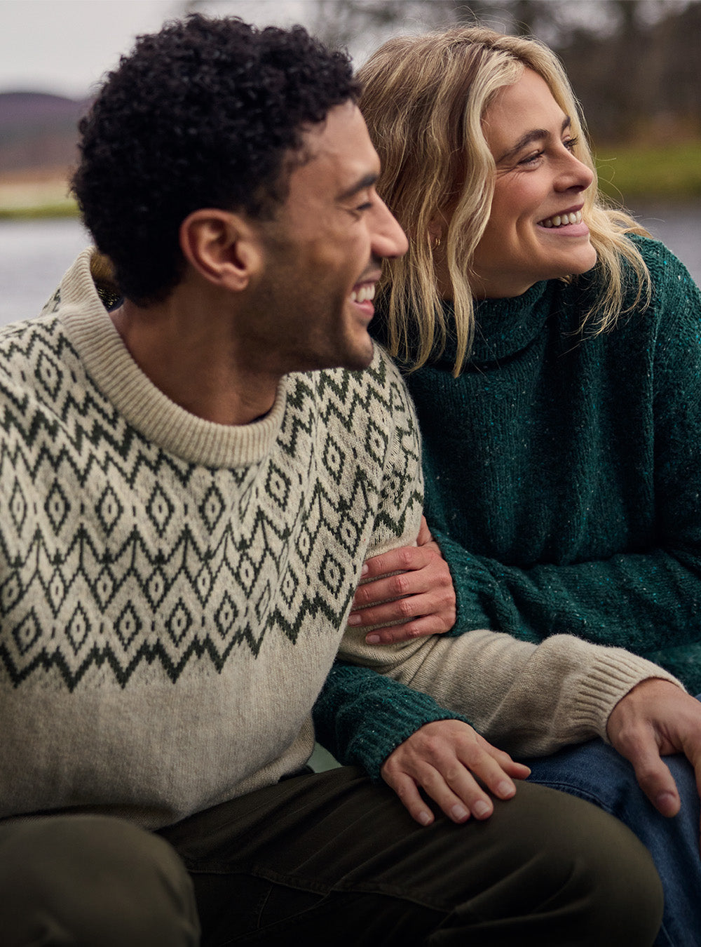 Man and woman smiling outdoors in cream and green knit sweaters.