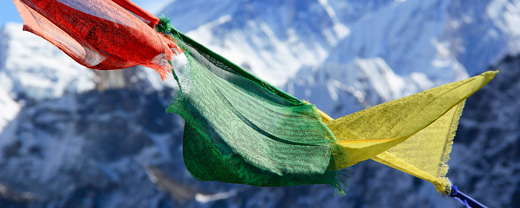 A row of red, green and yellow prayer flags blow in the wind in front of a mountain in Nepal. The mountain is covered in rocks and snow covers the peaks