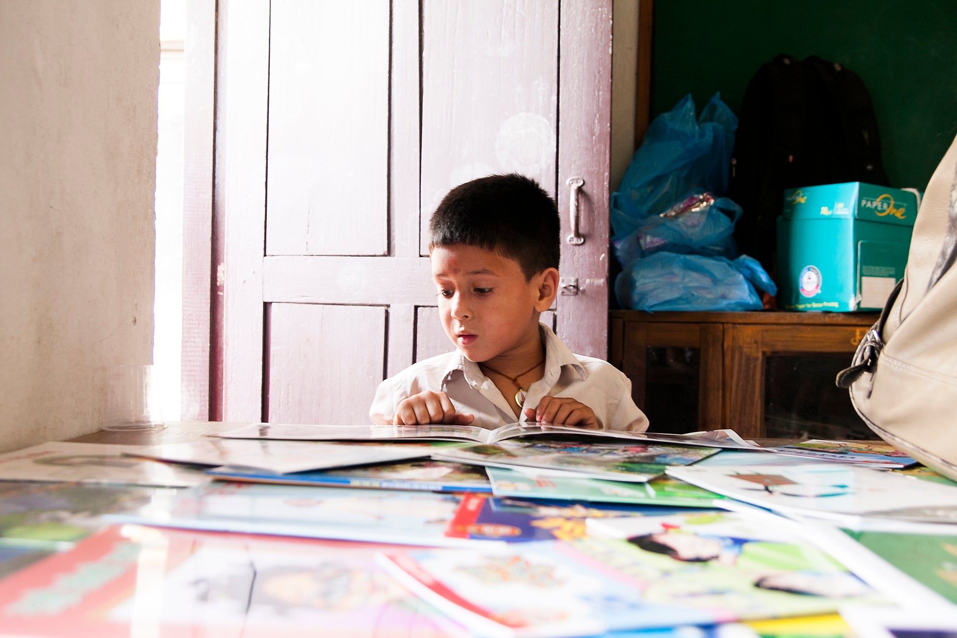 A Nepali child reading a picture book at a table with various other books laying on the table.