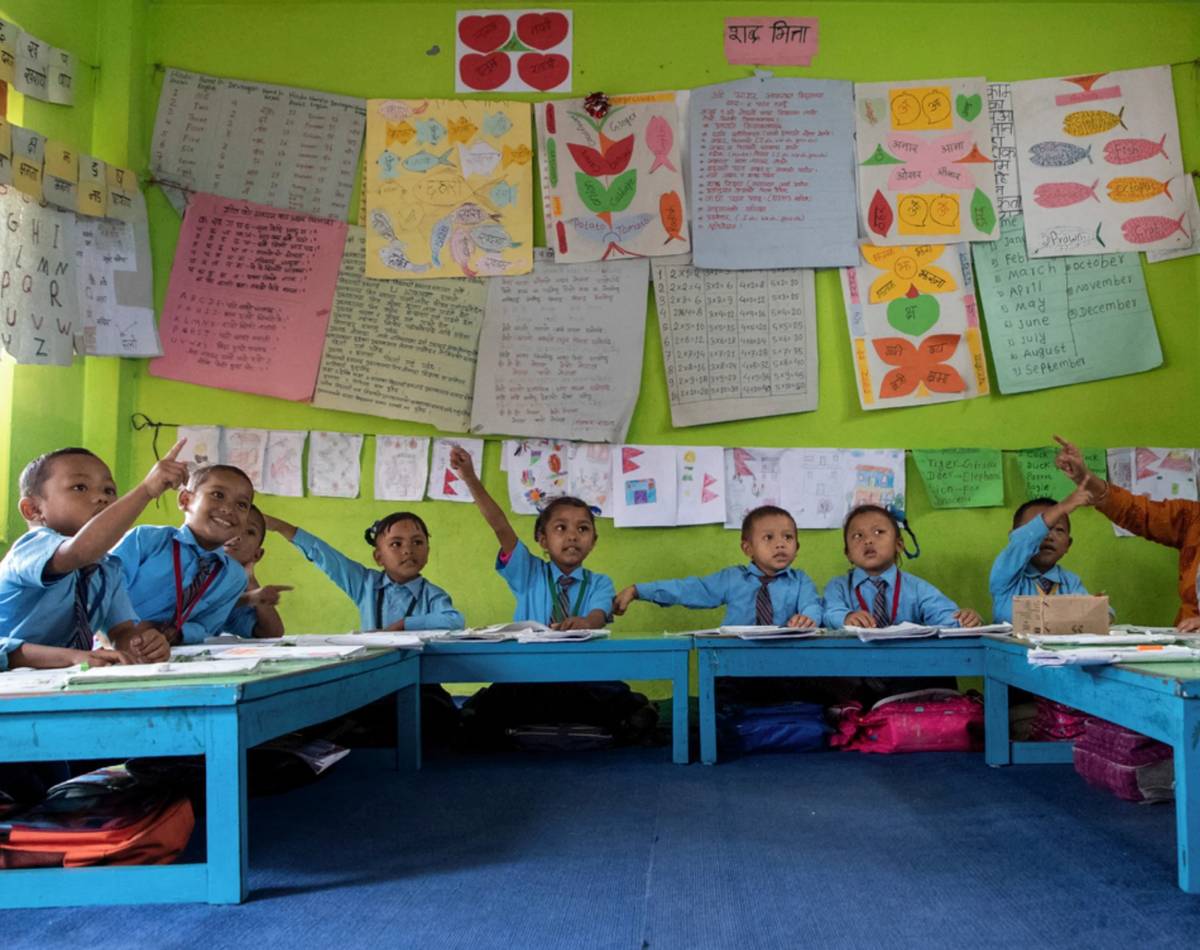 A group of smiling young children sit around a table in a Nepalese school, with their hands raised to answer questions. School art and work are pinned to the walls around them.
