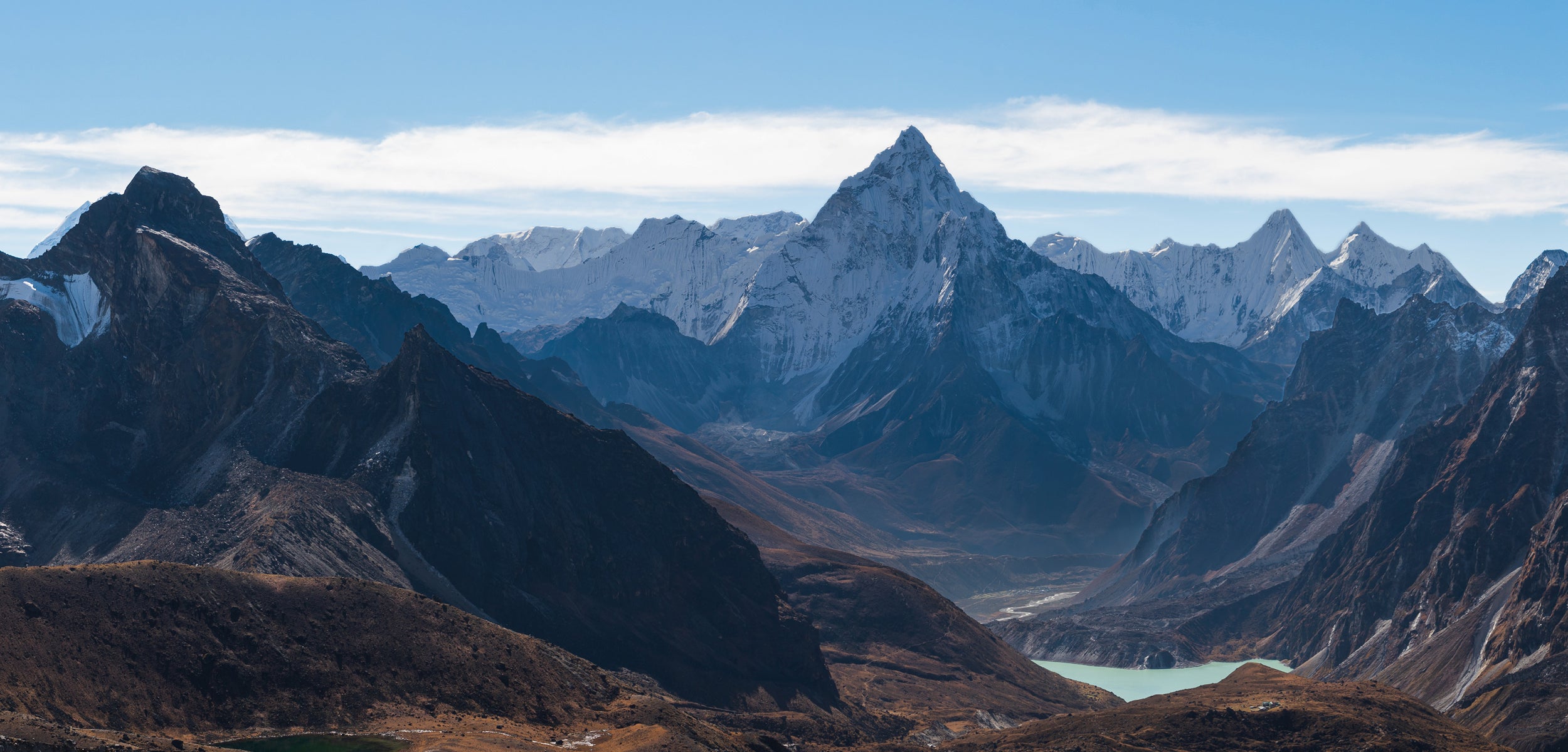 An image a mountain range in Nepal. In the foreground there are lower hills and a lake. In the background, there are tall snow capped mountains and clear blue sky with small clouds in the distance.
