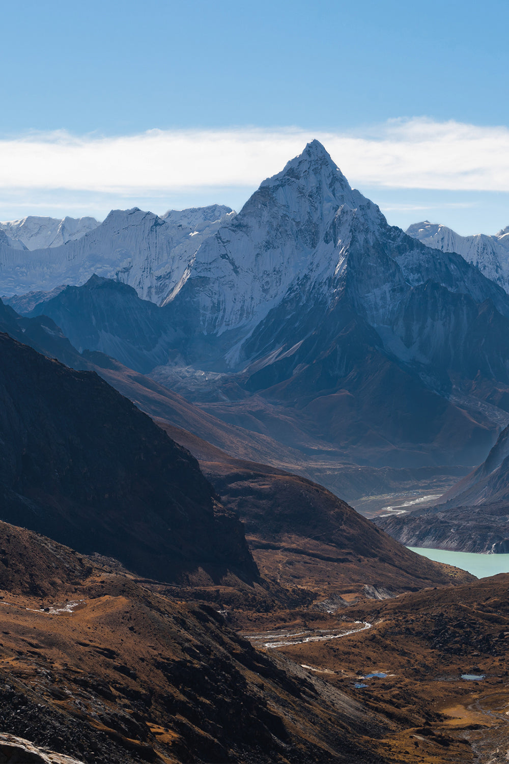 An image a mountain range in Nepal. In the foreground there are lower hills and a lake. In the background, there are tall snow capped mountains and clear blue sky with small clouds in the distance.