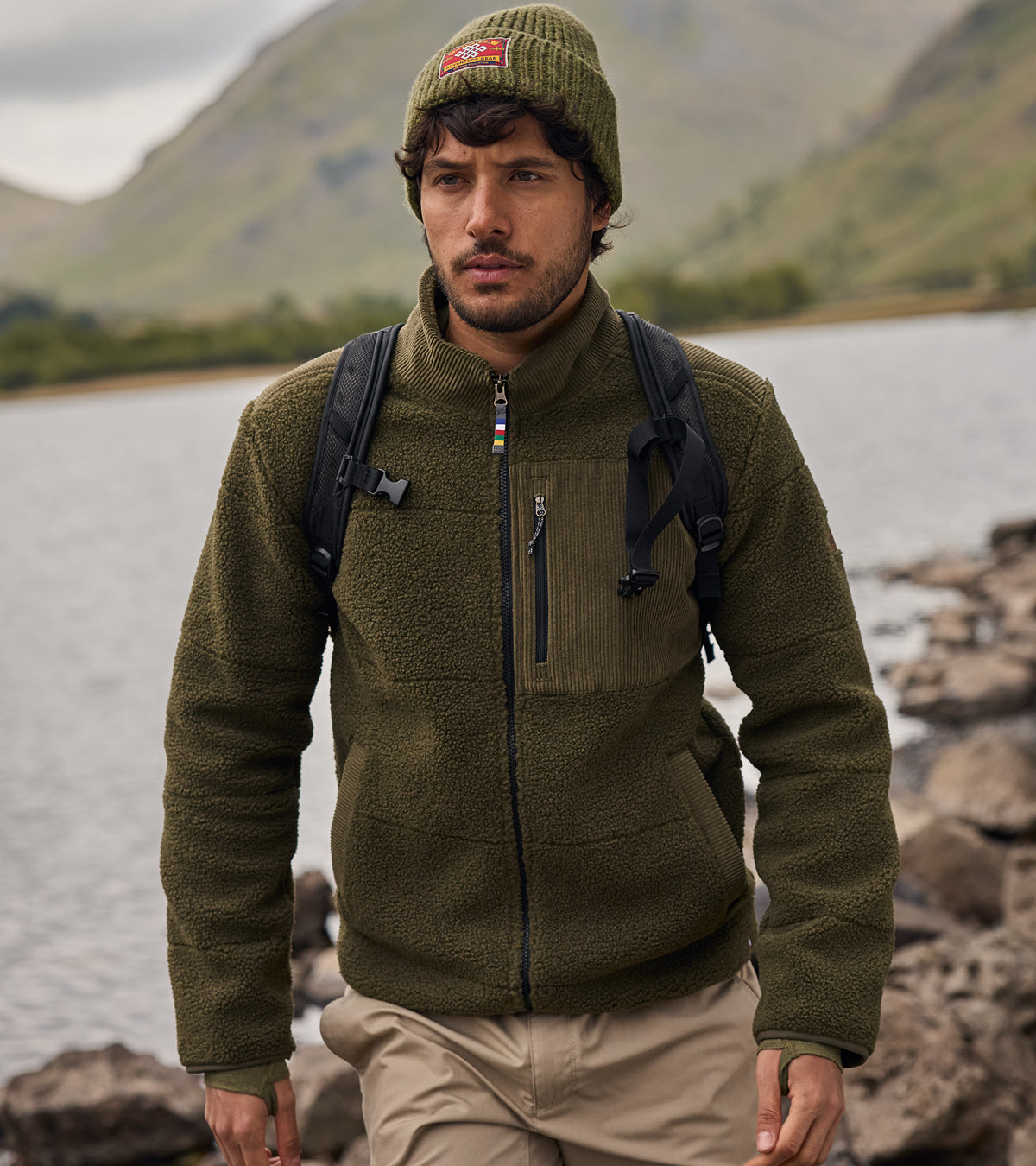 Man is walkin by a lake with tall, green hills in the background. He is wearing a green Sherpa Adventure Gear fleece jacket and a green beanie with a red patch on the front. He is carrying a black backpack with two straps visible on his shoulders.