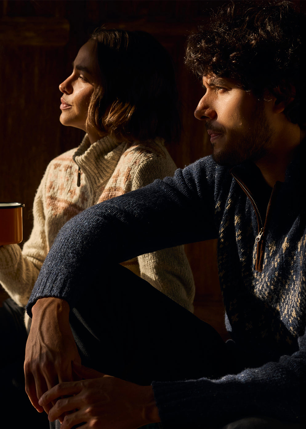 A man and a women sitting together in a warm, dimly lit wooden room holding mugs. The light is coming through a window and illuminating their faces. The women is wearing a cream and brown zip up cardigan. The man is wearing a navy blue quarter zip pullover.