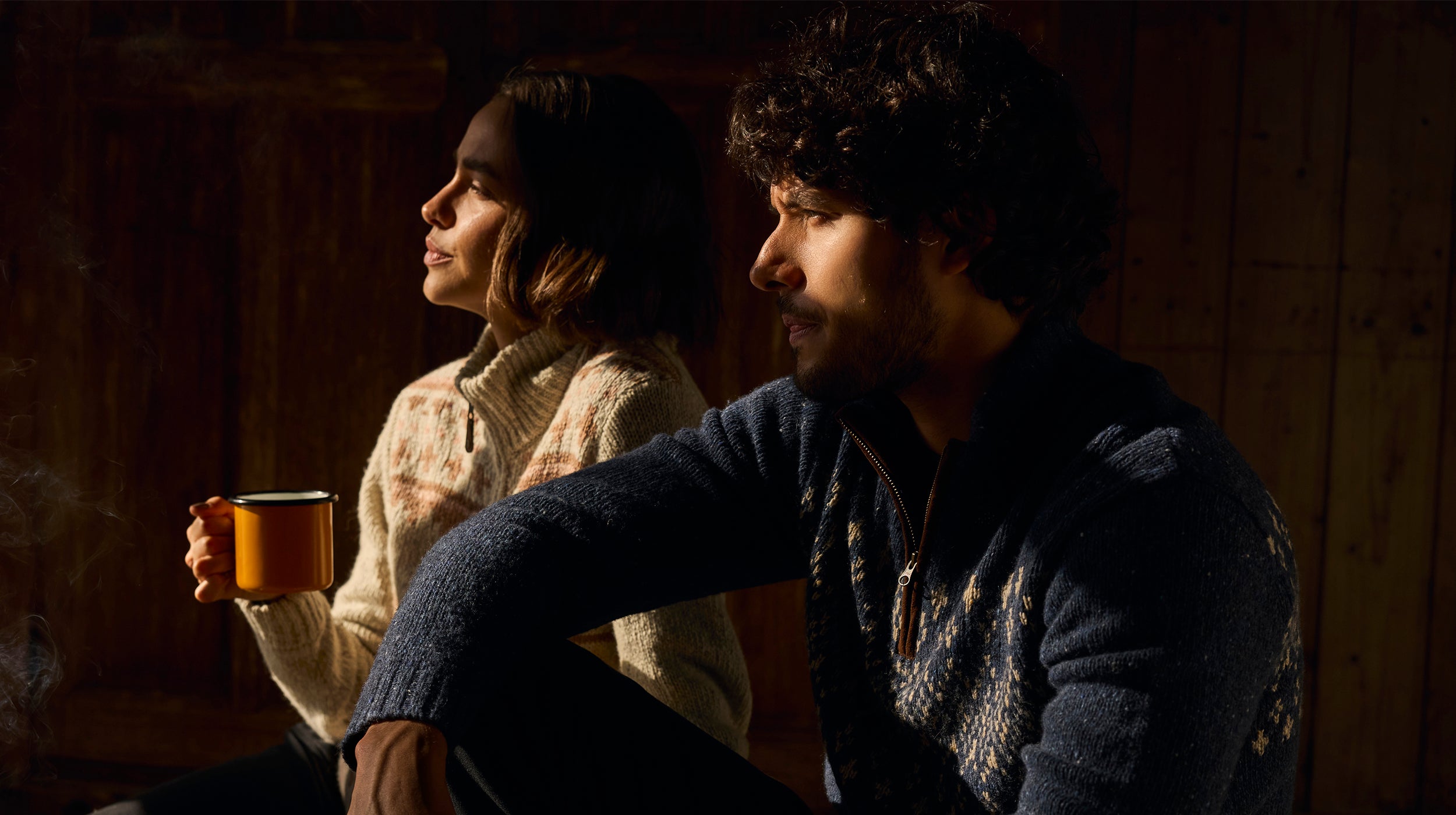 A man and a women sitting together in a warm, dimly lit wooden room holding mugs. The light is coming through a window and illuminating their faces. The women is wearing a cream and brown zip up cardigan. The man is wearing a navy blue quarter zip pullover.