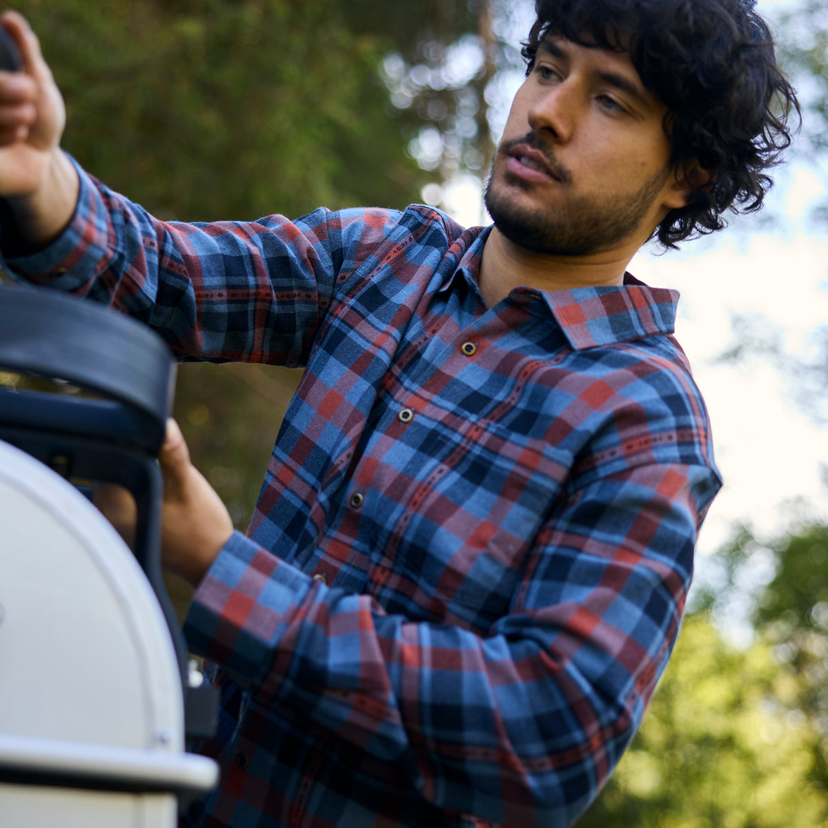 A close up of a man in a blue and red plaid shirt climbing on the back of a jeep. He is reaching for the car box on the roof.