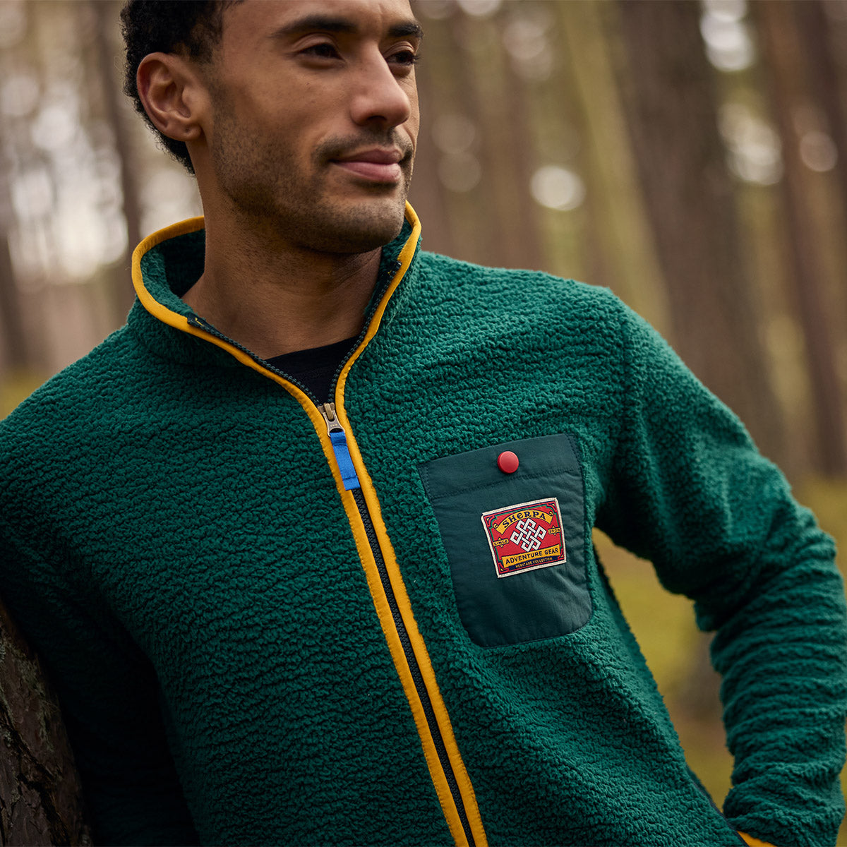 Man in a green fleece jacket with yellow trim leaning against a tree in a forest.