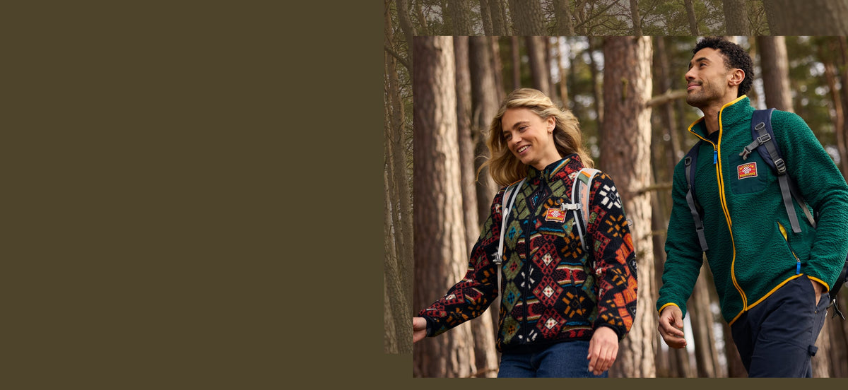 Two people hiking in a pine forest, wearing patterned and green fleece jackets.
