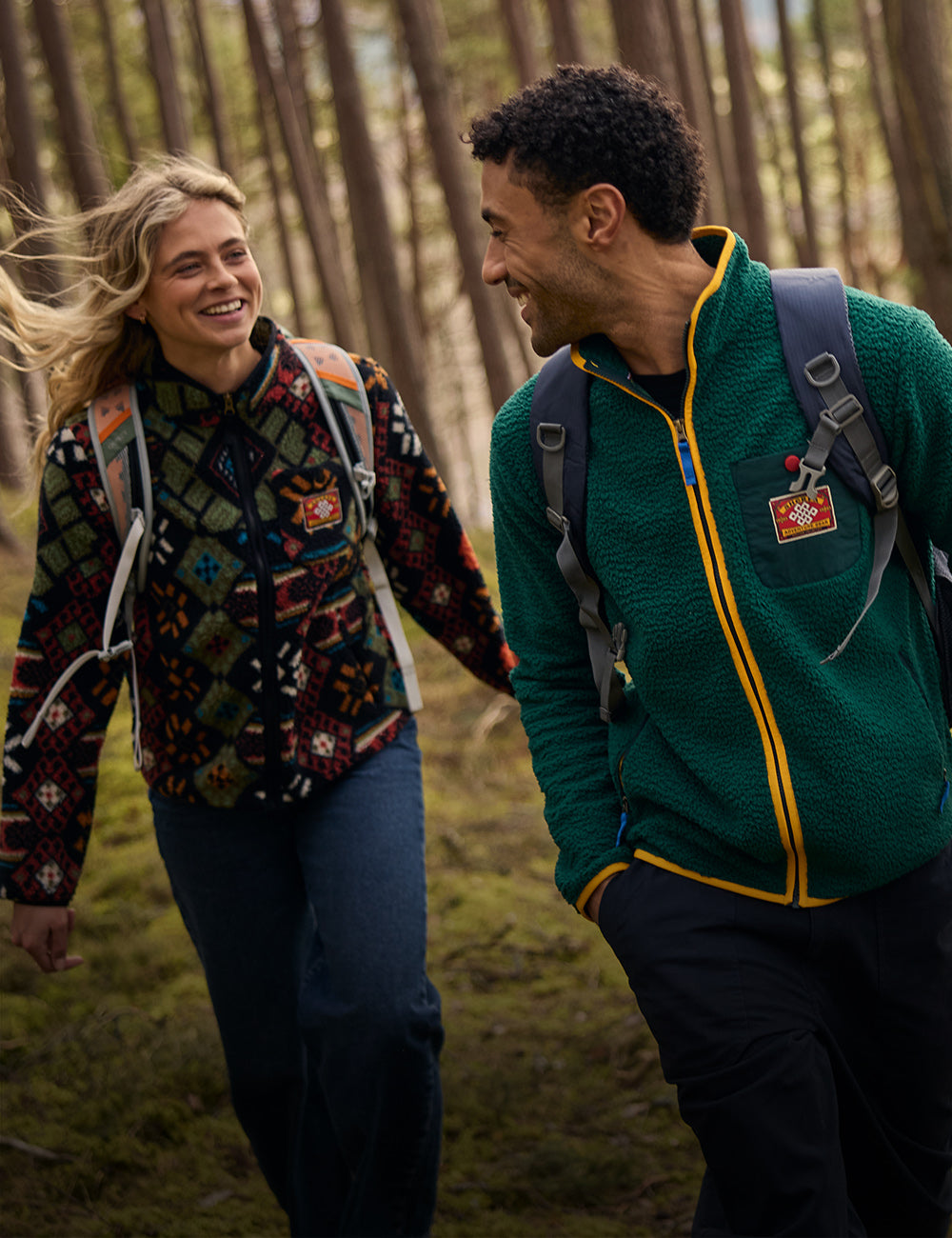 A man and woman in a forest, both wearing patterned fleece jackets with red and yellow logo patches