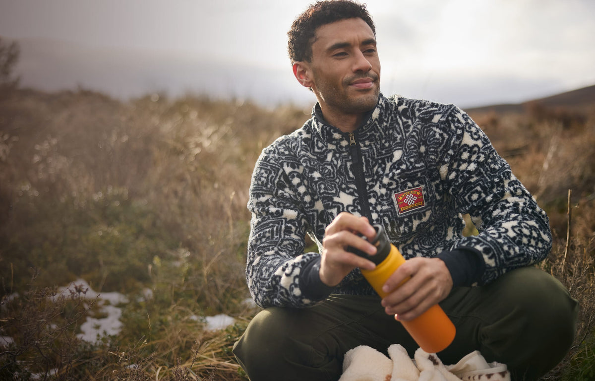 A man kneeling outdoors in a grassy field, wearing a black and white patterned fleece jacket.
