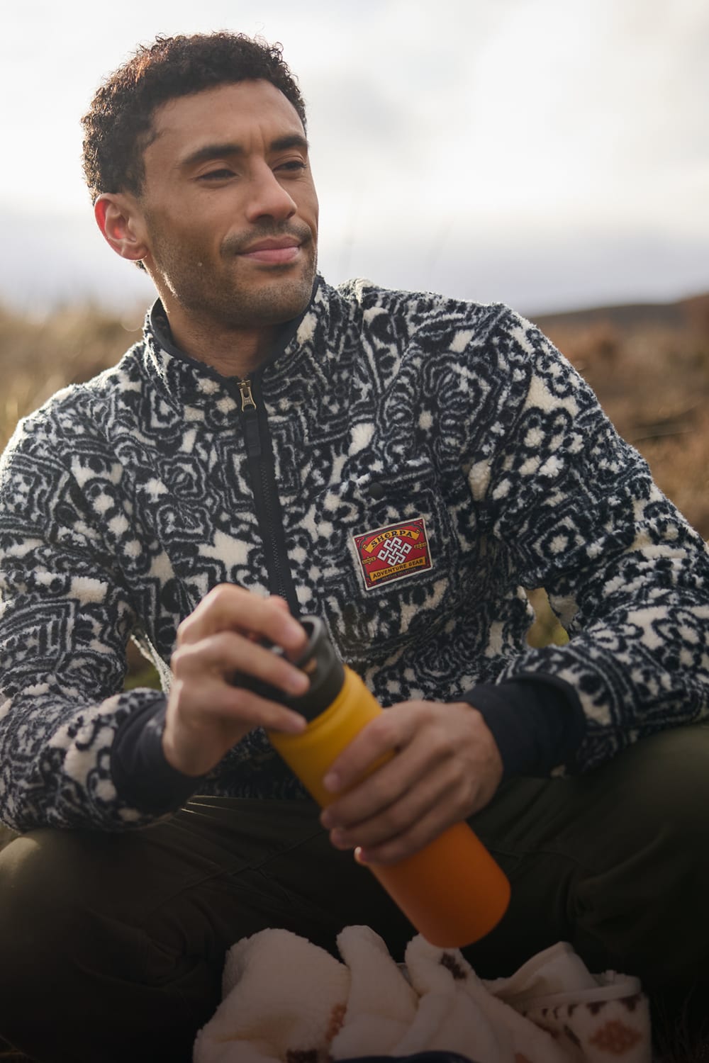 Man wearing a black and white patterned fleece jacket with a logo patch, holding an orange water bottle outdoors.