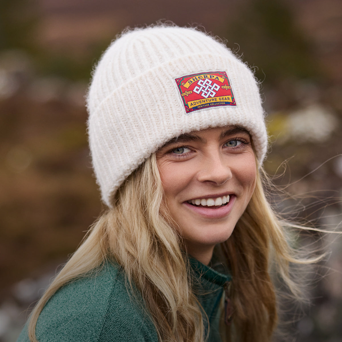 Smiling woman in a cream knit beanie with a colourful embroidered patch outdoors.