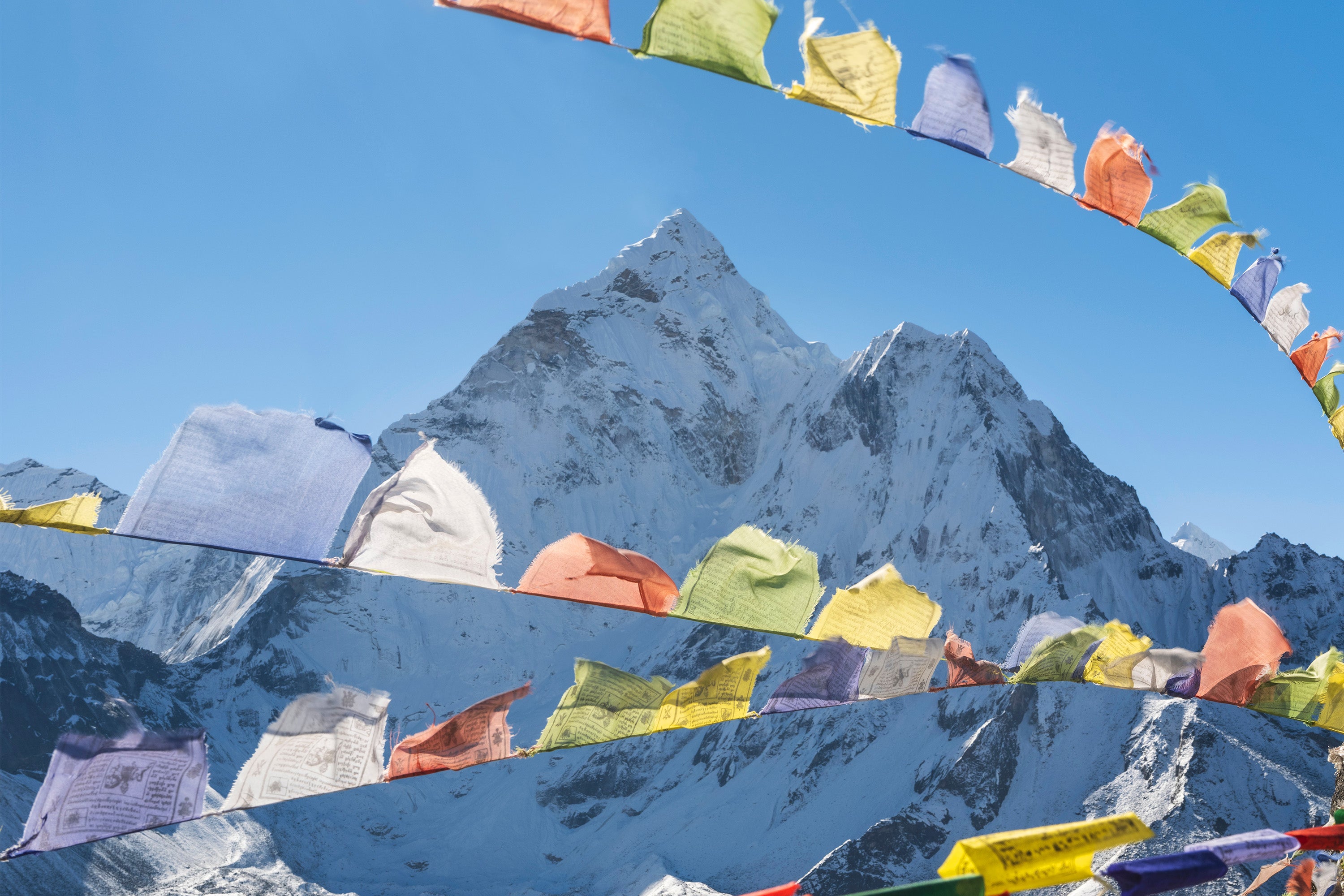 Four rows of prayer flags are fluttering with Mount Everest in the background. The flags are small squares of cloth with Nepali writing and symbols on both sides. The flags are brightly coloured with vibrant reds, whites, blues, yellows and greens