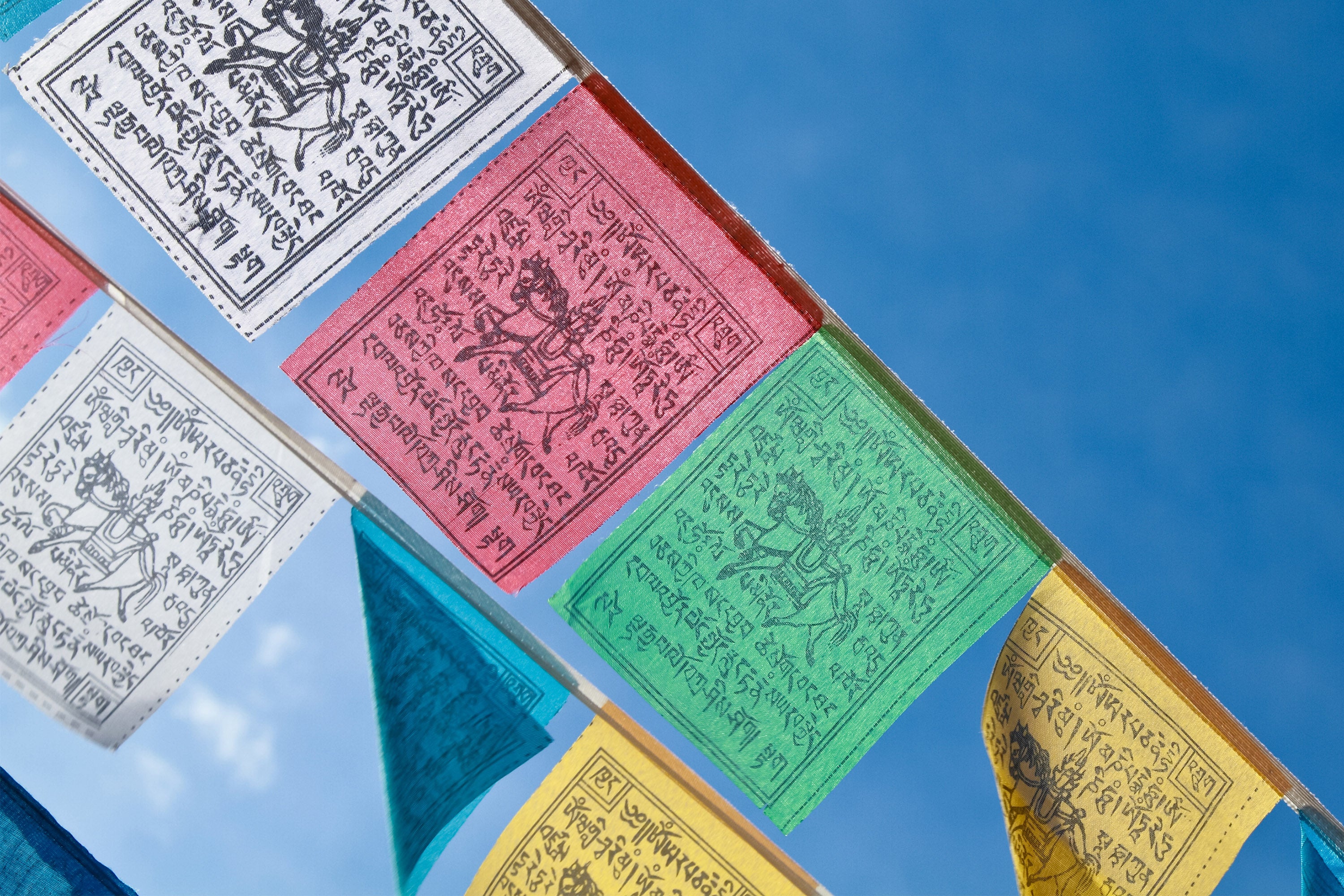 A close up of Nepali prayer flags against a blue, cloudless sky. The flags dyed in vibrant colours: yellow, green, red, white and blue. Each flayer has a depiction of a window horse surrounded by Nepali lettering.