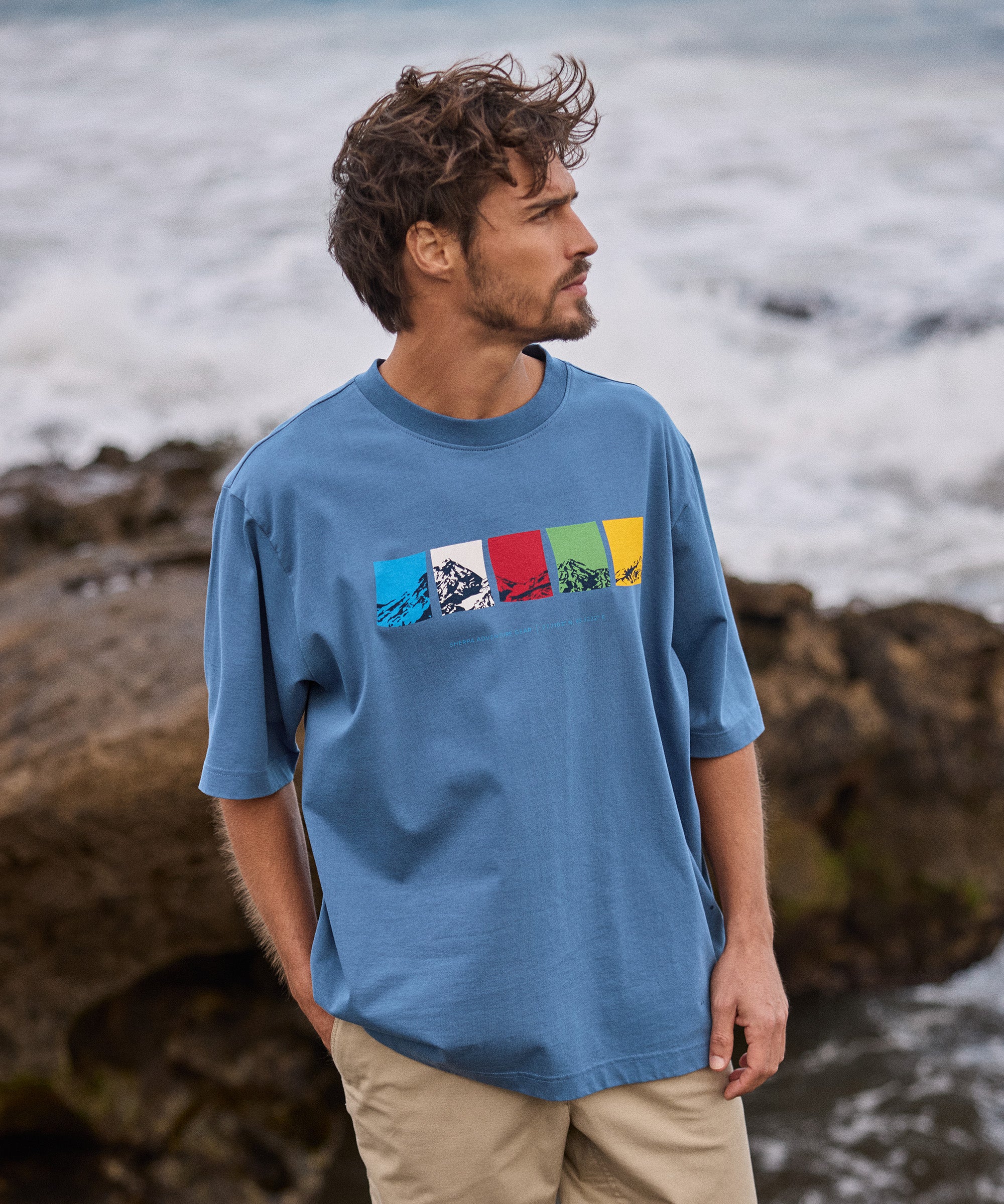 Man wearing a blue t-shirt with colorful graphic design by the ocean. The graphic depicts the 5 colours of the Nepali prayer flags: Blue, white, red, green and yellow.