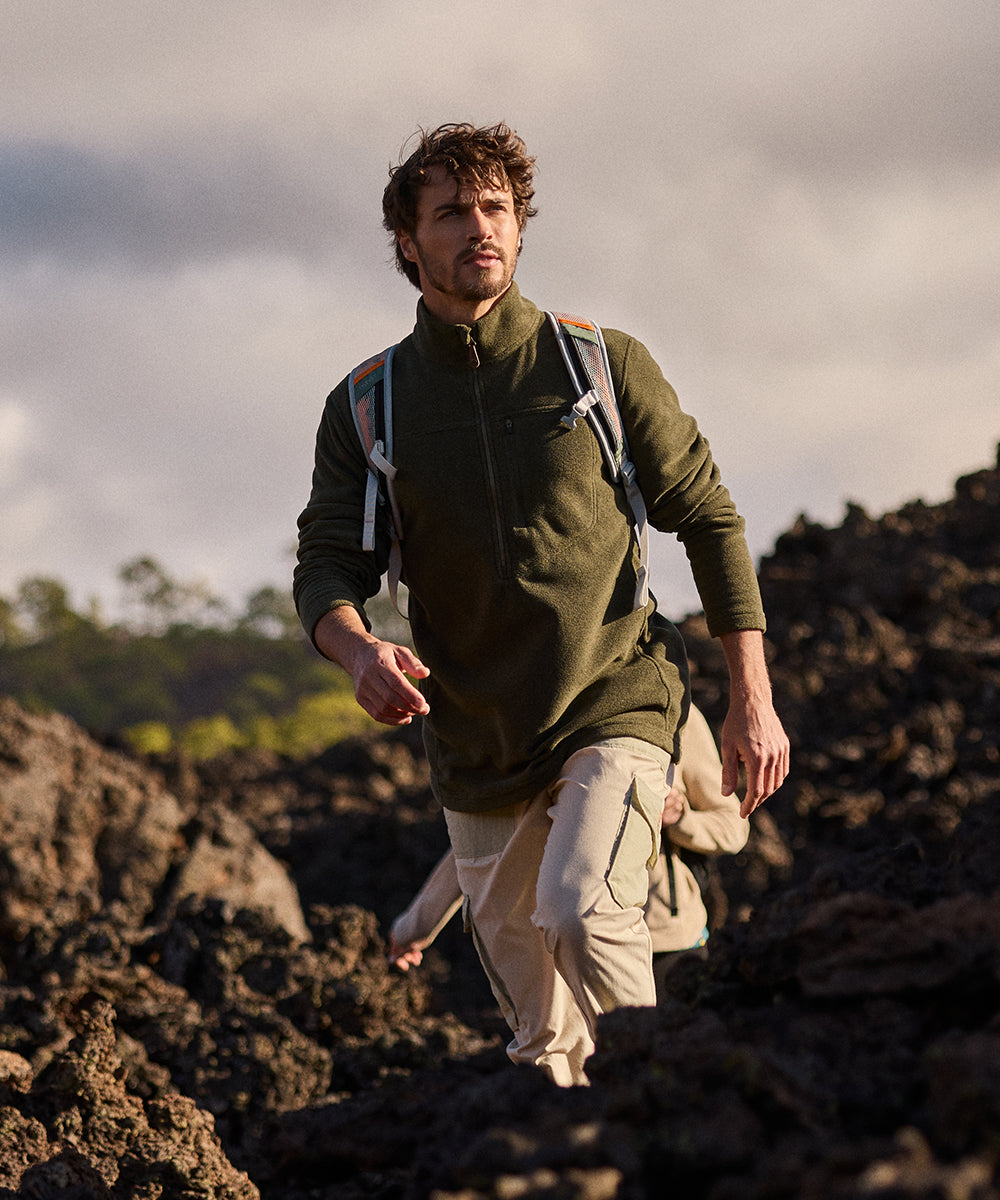 Man hiking on a rocky landscape with a backpack. He is wearing a green, half zip fleece and cream cargo trousers.