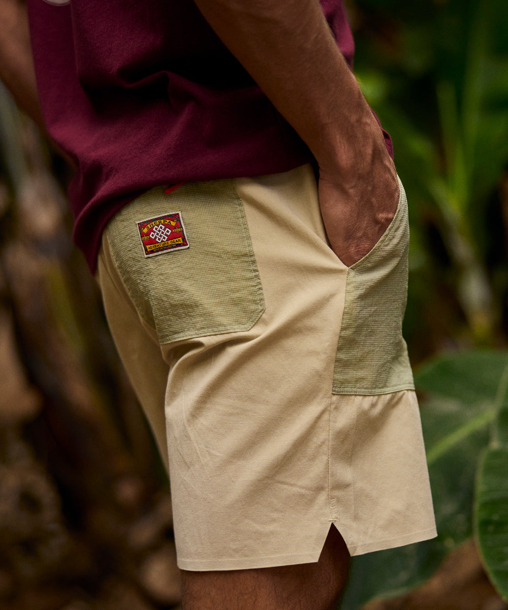 A man wearing a pair of beige shorts and a red T-shirt. The shorts have green pockets on the front and back with a small, red "Sherpa Adventure Gear" patch on the back pocket. He is standing with his hands in his pockets against a forest background.