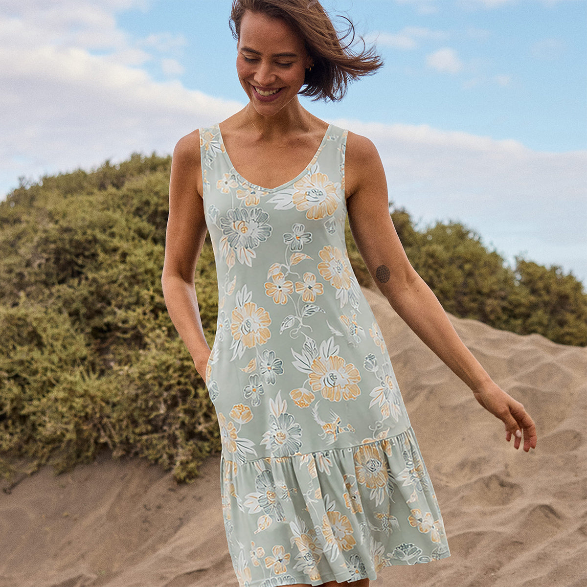 A woman wearing a green floral dress standing on a sandy landscape with greenery. She is smiling looking down at the golden sand with her hand in her dress pocket.