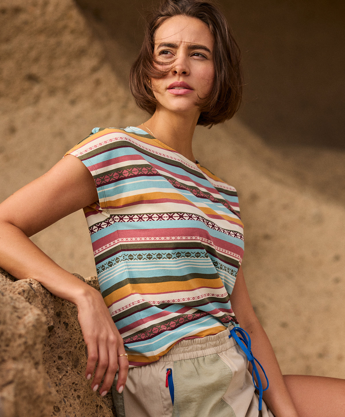 A woman wearing a colorful striped T-shirt against a yellow, sandstone wall. The T-Shirt is paired with a beige skort with green pockets and a blue drawcord. She is standing, resting her arm on the rock face, looking out into the distance.