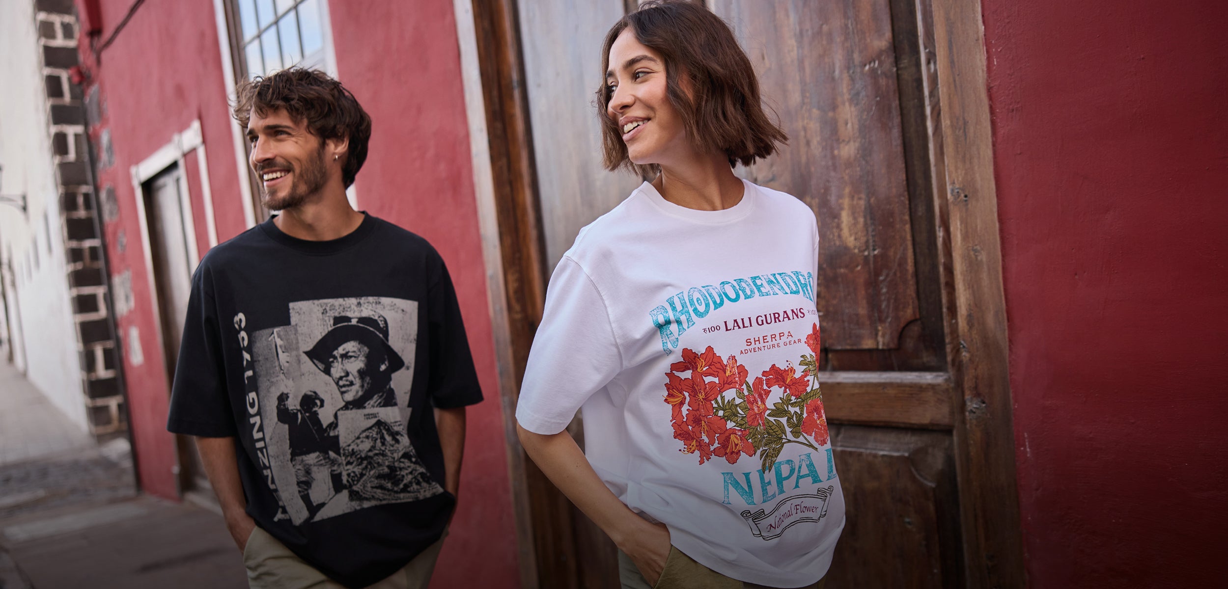 A man and a woman walking down a Spanish street. The man is wearing a black T-Shirt depicting Tenzing Norgay. The woman is wearing a white T-Shirt depicting a red rhododendron flower. They are smiling and have their hands in their pocket.