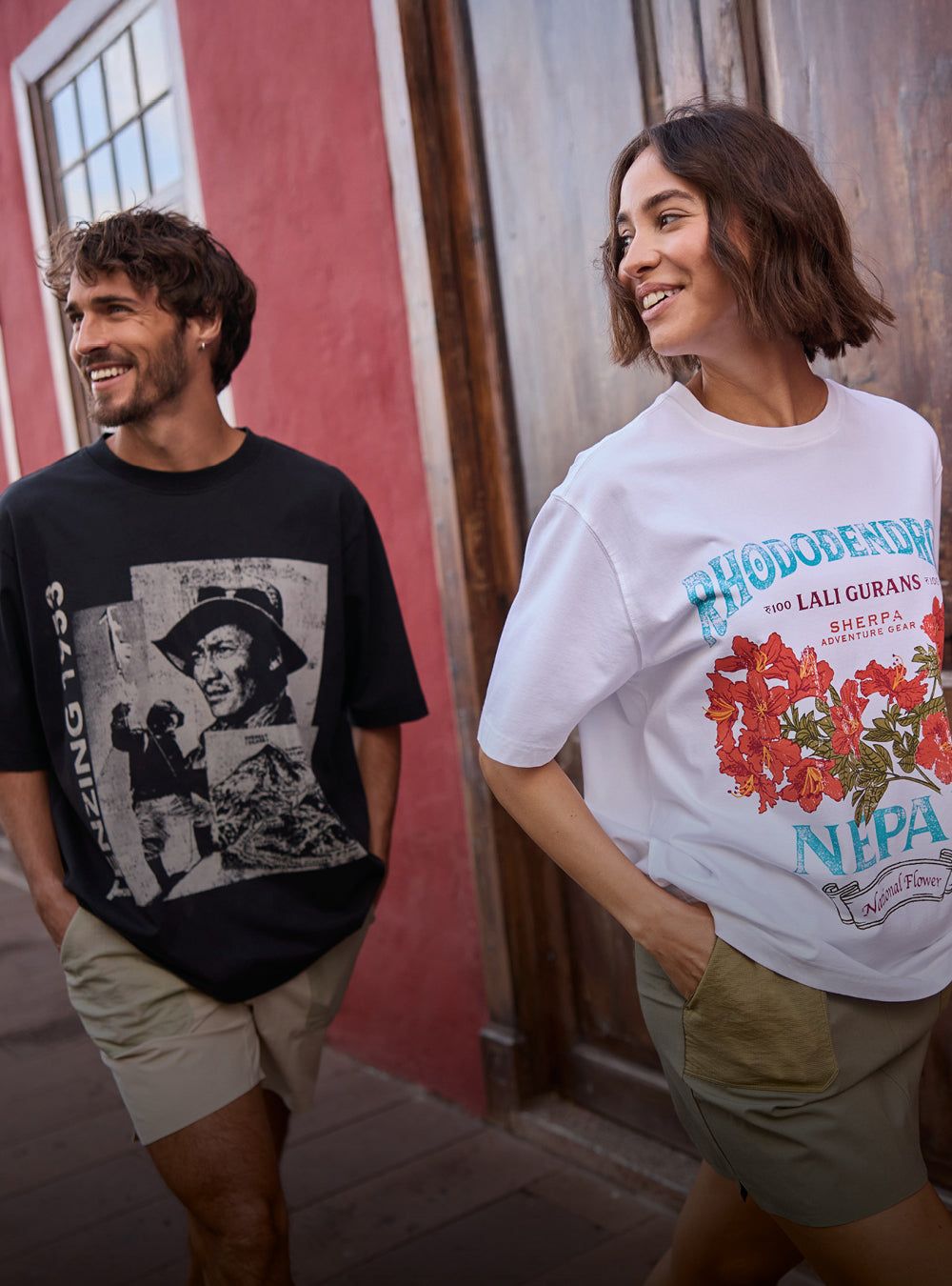 A man and a woman walking down a Spanish street. The man is wearing a black T-Shirt depicting Tenzing Norgay. The woman is wearing a white T-Shirt depicting a red rhododendron flower. They are smiling and have their hands in their pocket.