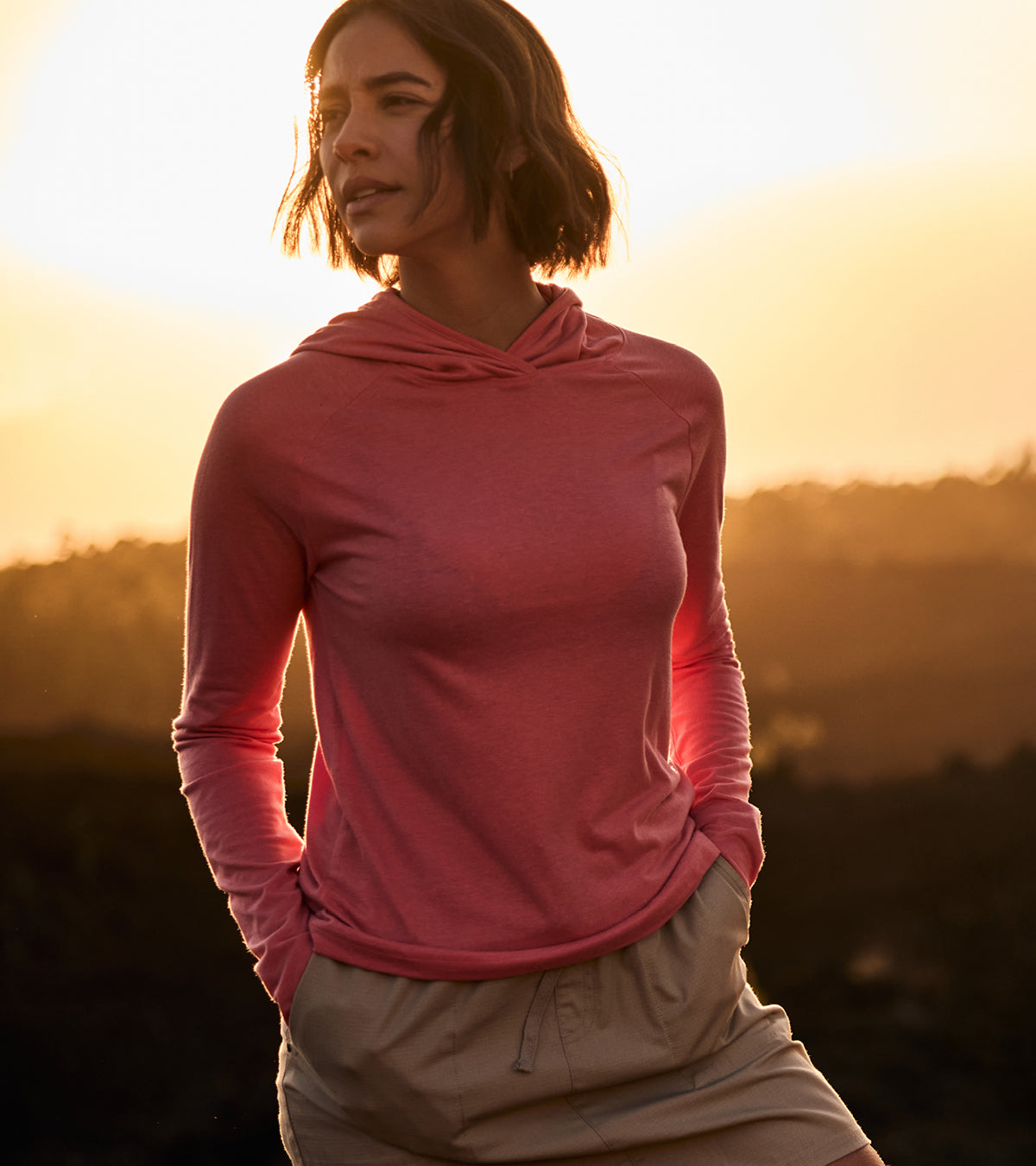 A woman wearing a lightweight pink long-sleeve top with a hood against a sunset backdrop