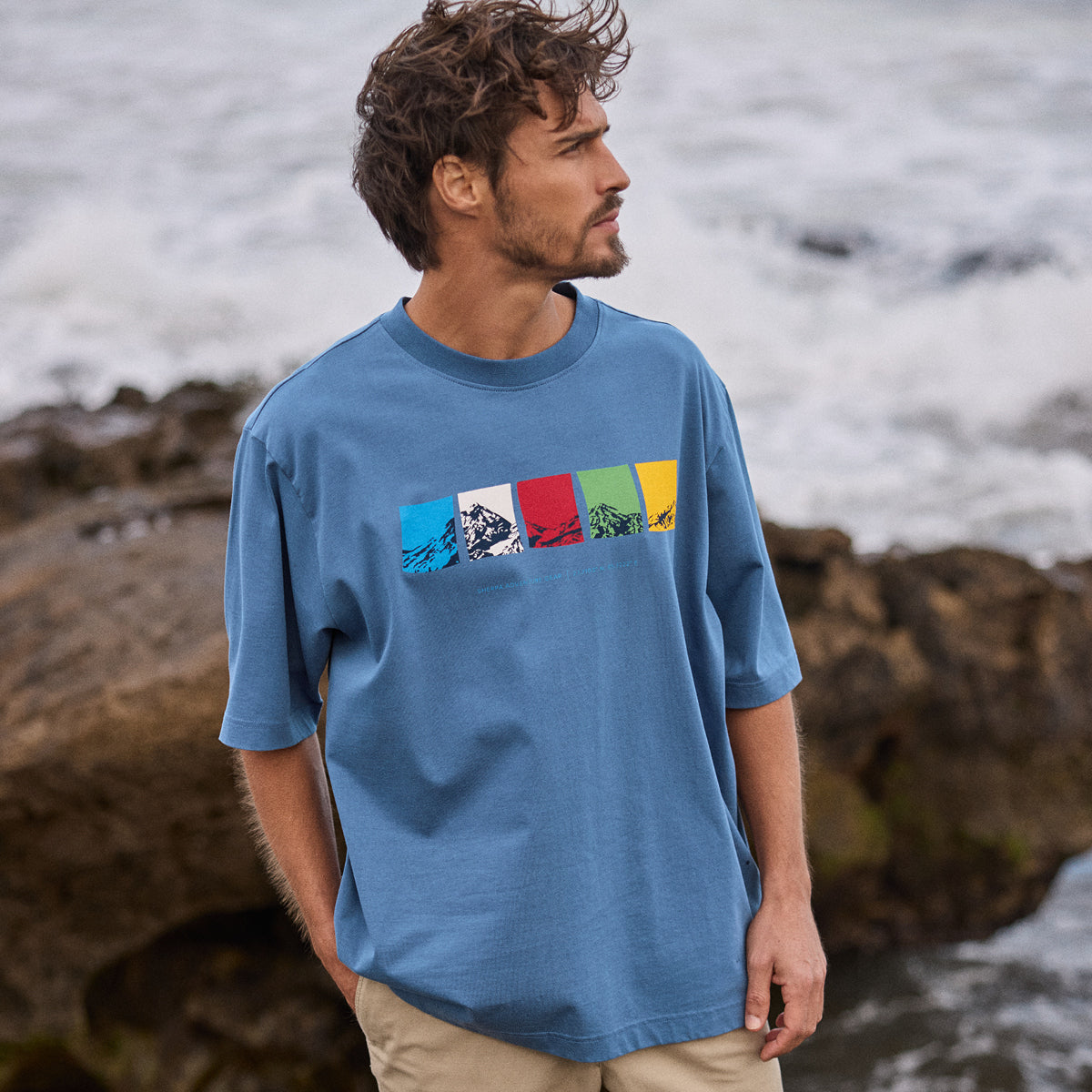 Man wearing a blue t-shirt with colorful Nepal prayer flag graphic by a rocky shoreline