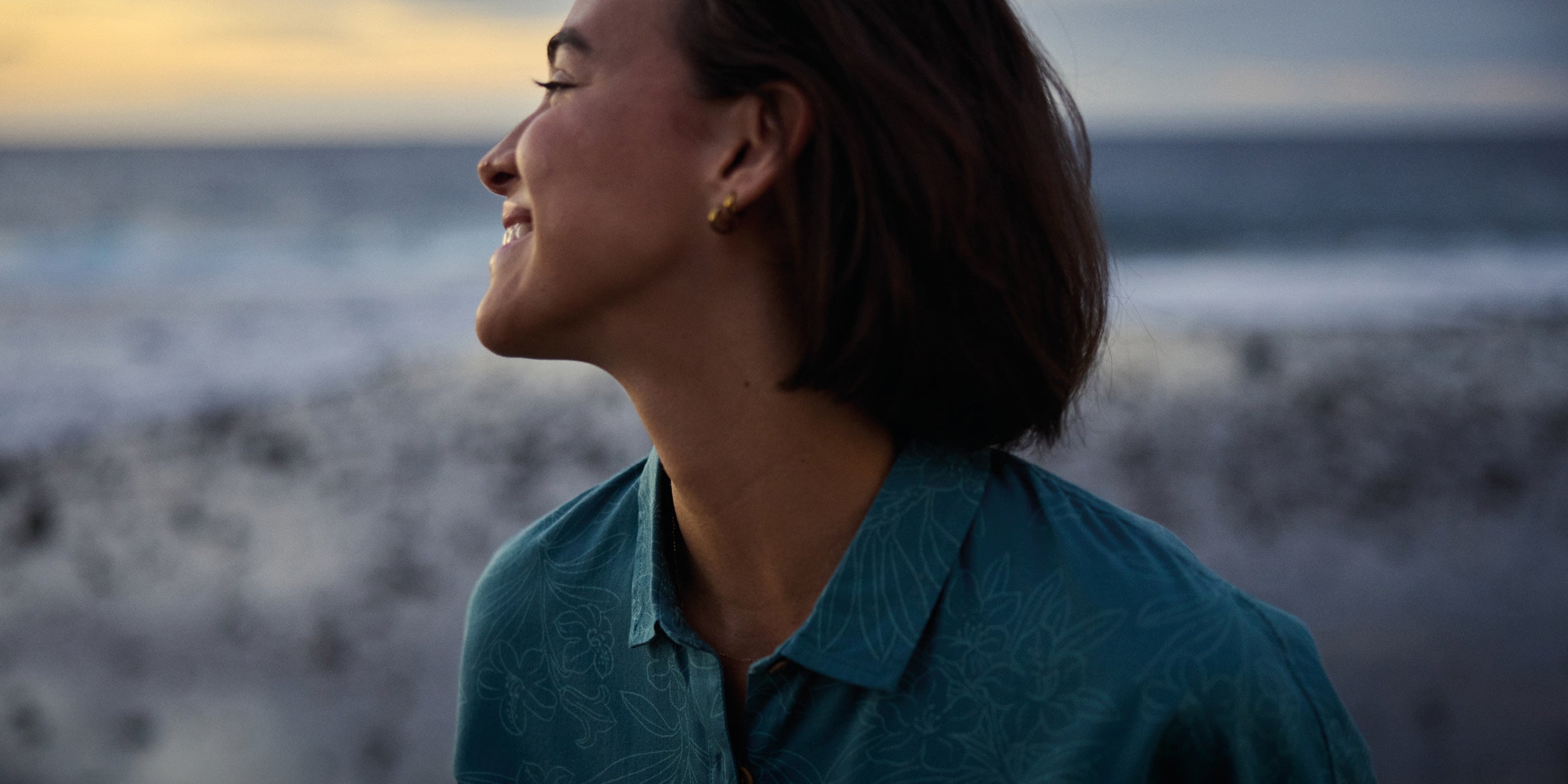 A close up of a woman in teal shirt standing on a beach at sunset. She is looking over her should smiling at the sunset.