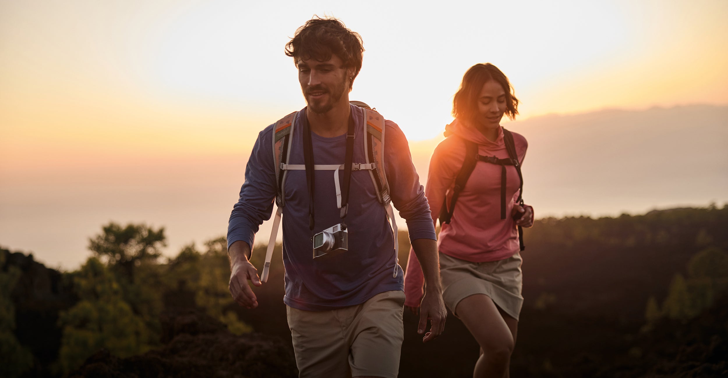 A man and a woman hiking on a hill at sunset. They have reached the crest of the hill and tree tops are visible around them. The man is wearing a blue long sleeve top, beige shorts, a backpack and a camera around his neck. The woman is slightly behind him, wearing a lightweight pink hoodie with a beige skort and a black backpack.