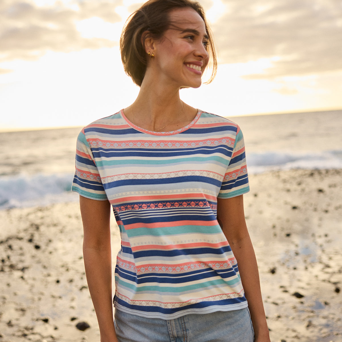 A woman standing on a pebble beach at sunset. She is smiling and looking to the side. She is wearing a pair of denim shorts and T-Shirt with horizontal stripes.