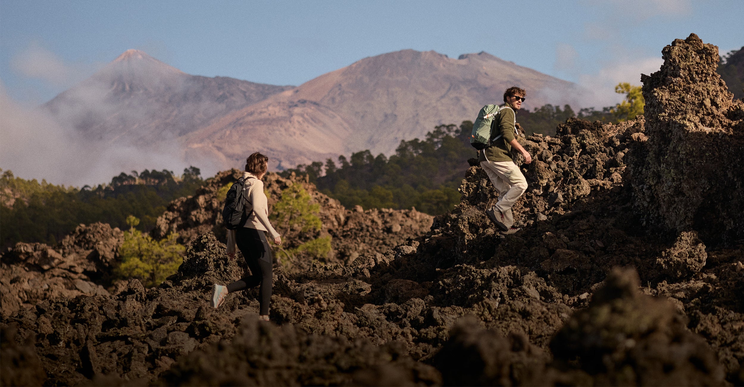 Two hikers on a rocky landscape with mountains in the background. The man is wearing a green fleece and white hiking trousers whilst carrying a green backpack. The woman is wearing a beige fleece and black legging and backpack.