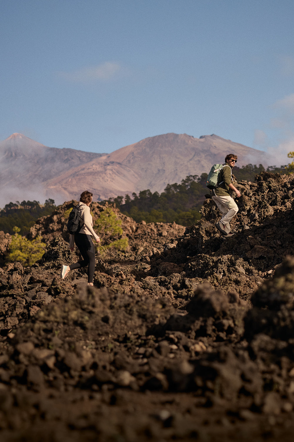 Two hikers on a rocky landscape with mountains in the background. The man is wearing a green fleece and white hiking trousers whilst carrying a green backpack. The woman is wearing a beige fleece and black legging and backpack.