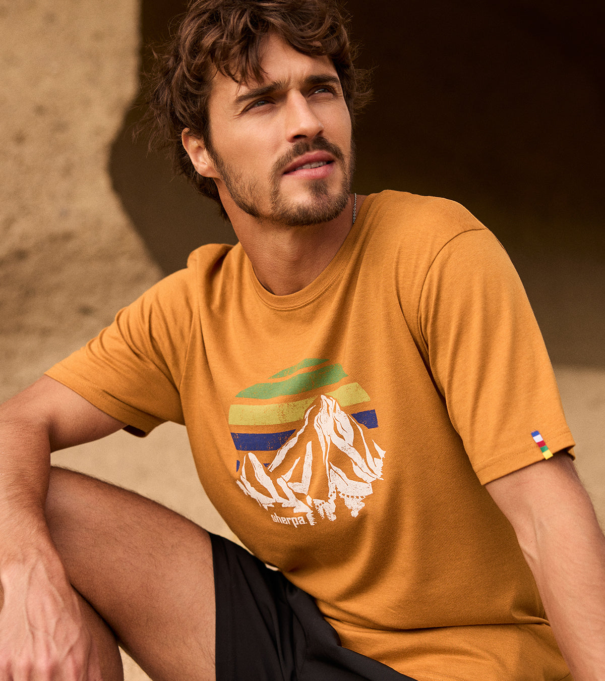 A man wearing a mustard yellow t-shirt with a graphic design, sitting against a natural sandstone wall. The graphic depicts a Nepali mountain peak and a sunset of greens and blue. The brand name "Sherpa" can also be seen on the graphic.