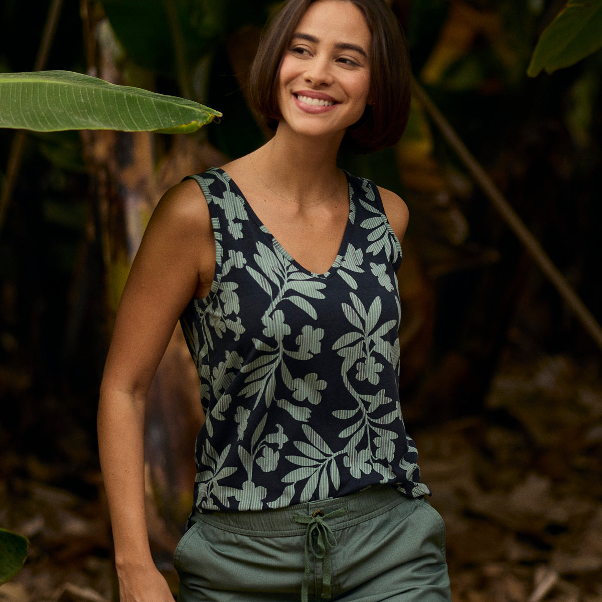 A woman wearing a floral tank top and green pants standing in a forest setting. She is walking through the forest smiling.